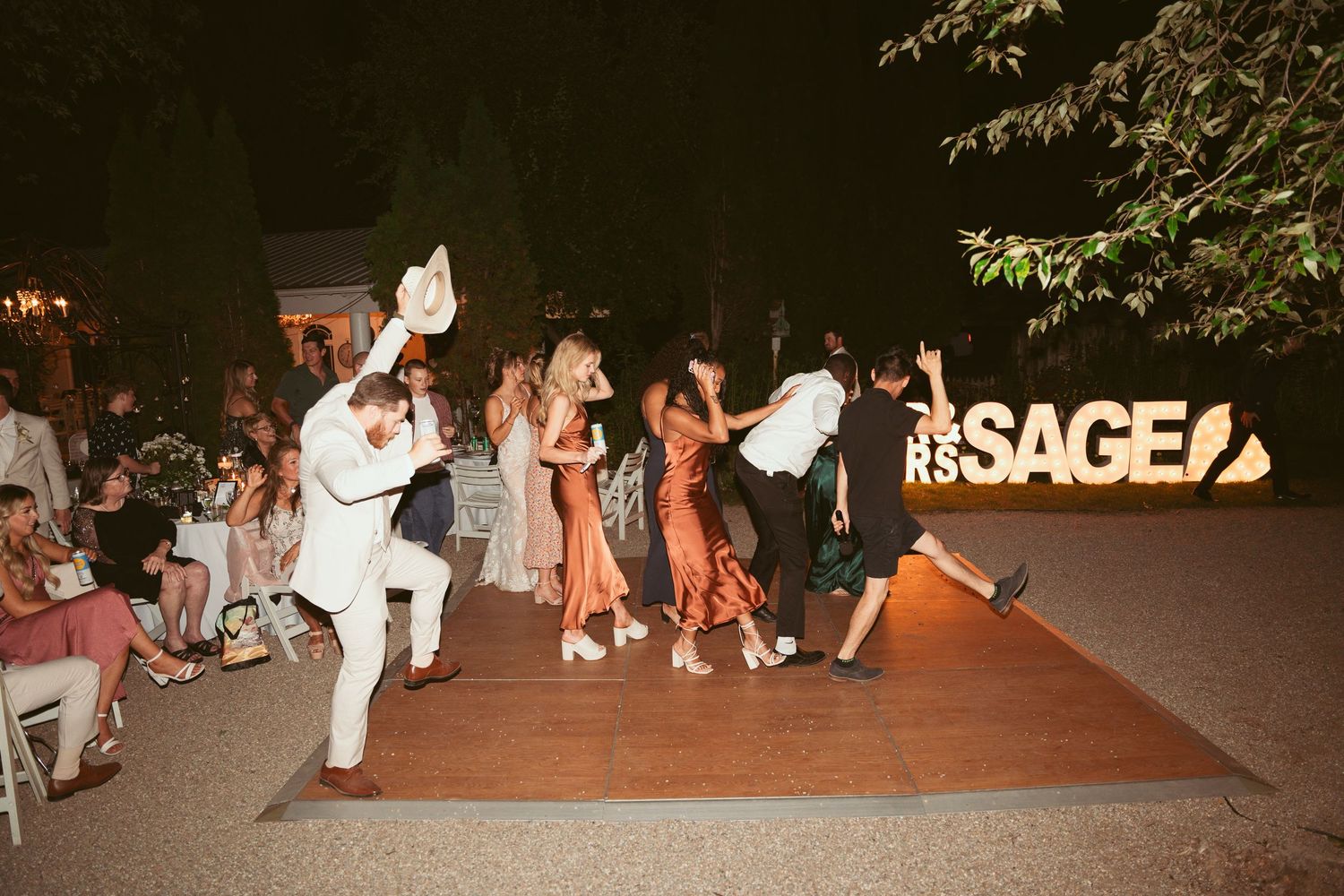 Wedding guests dancing and mingling on outdoor patio with lit SAGE sign.
