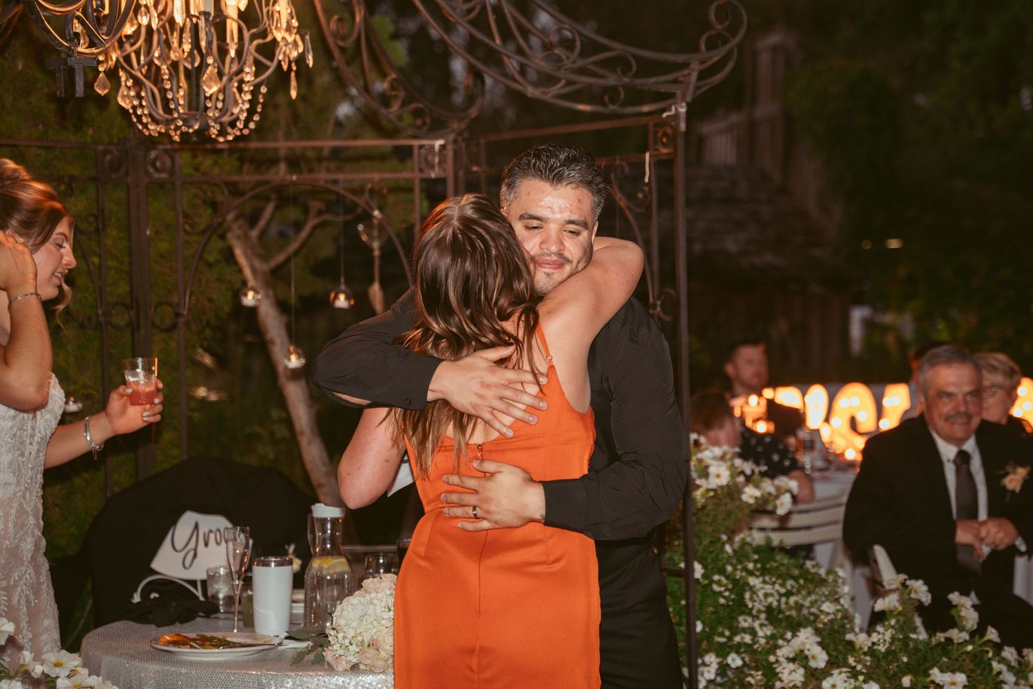 Two people share an emotional embrace during an evening reception under a chandelier.
