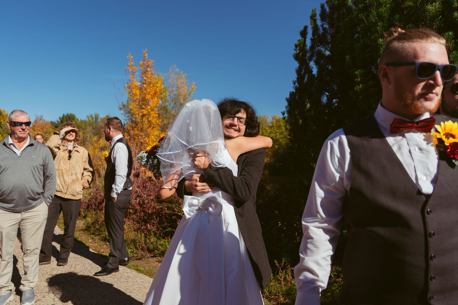 Wedding party members interact during casual outdoor photo session with fall foliage.