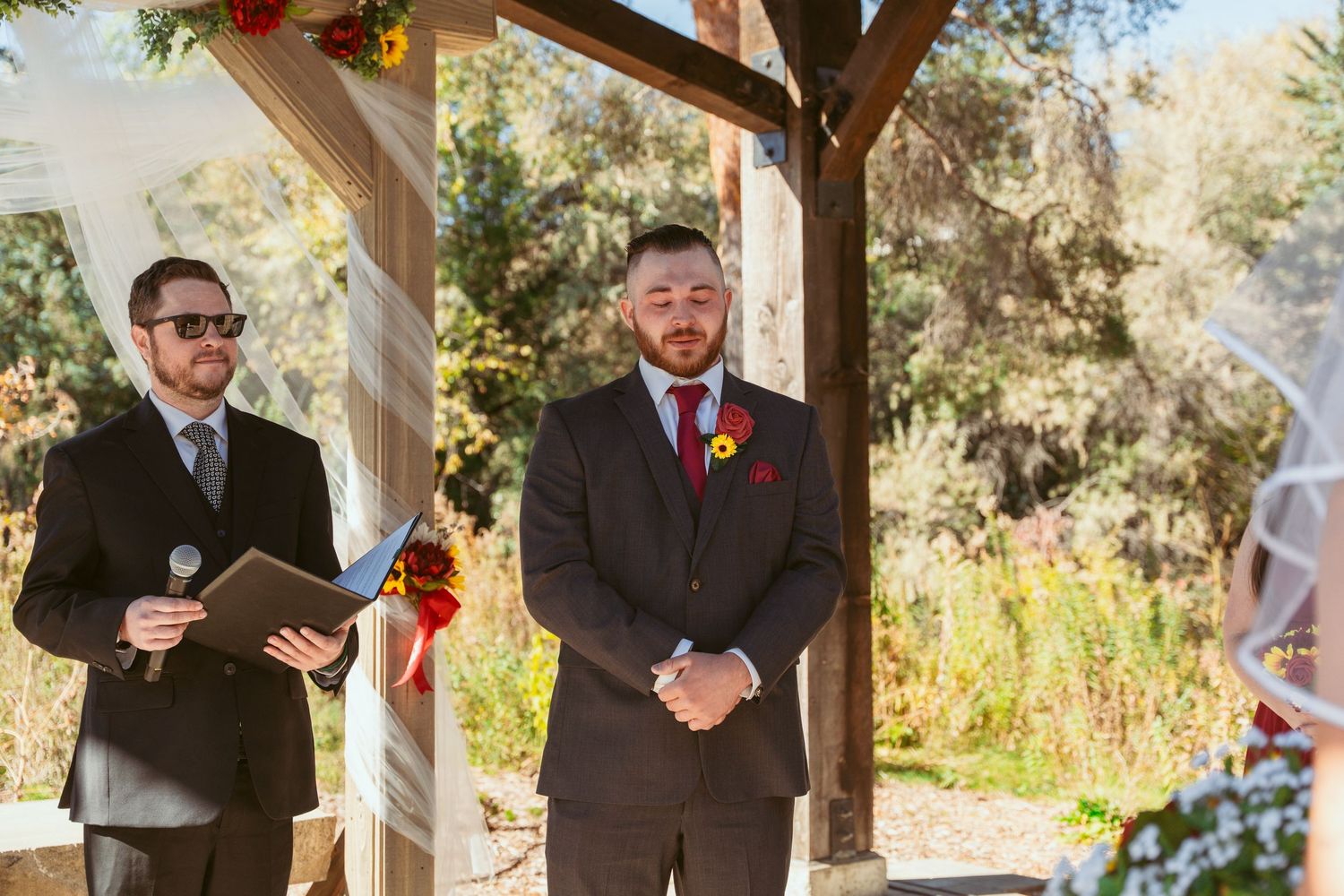 Wedding ceremony guests stand under wooden beams decorated with sunflowers and autumn foliage.