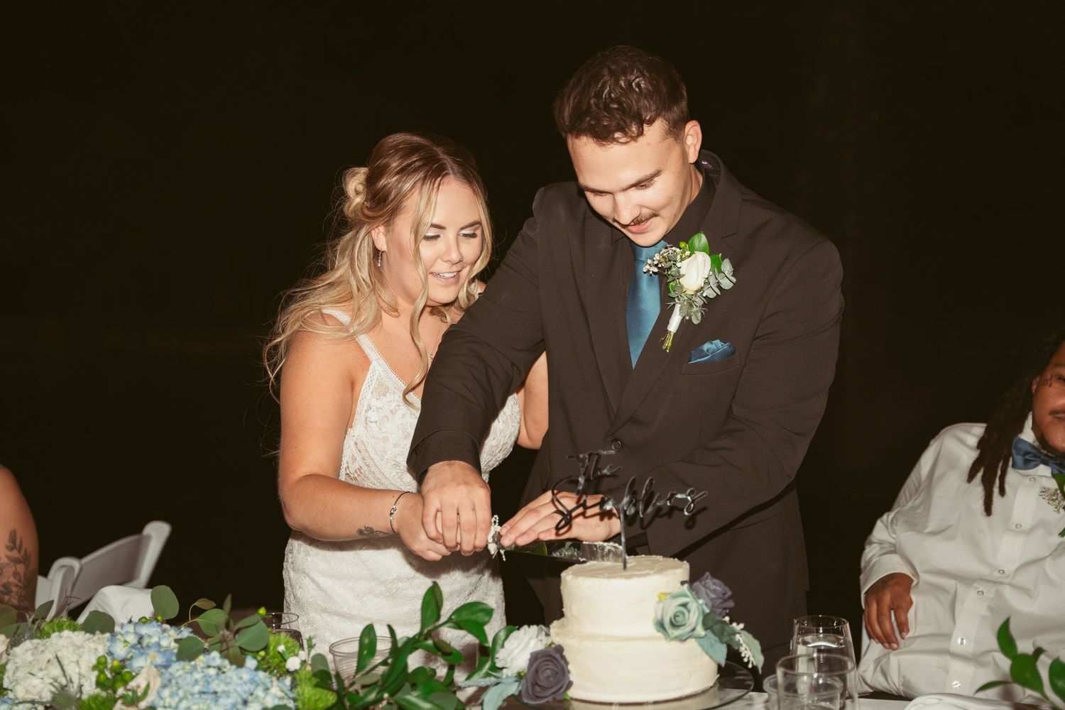 Newlyweds share a moment at their reception table decorated with blue hydrangeas and greenery.
