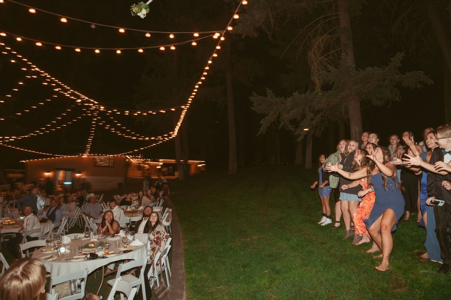 Wedding guests dance and celebrate under romantic string lights at an outdoor evening reception.