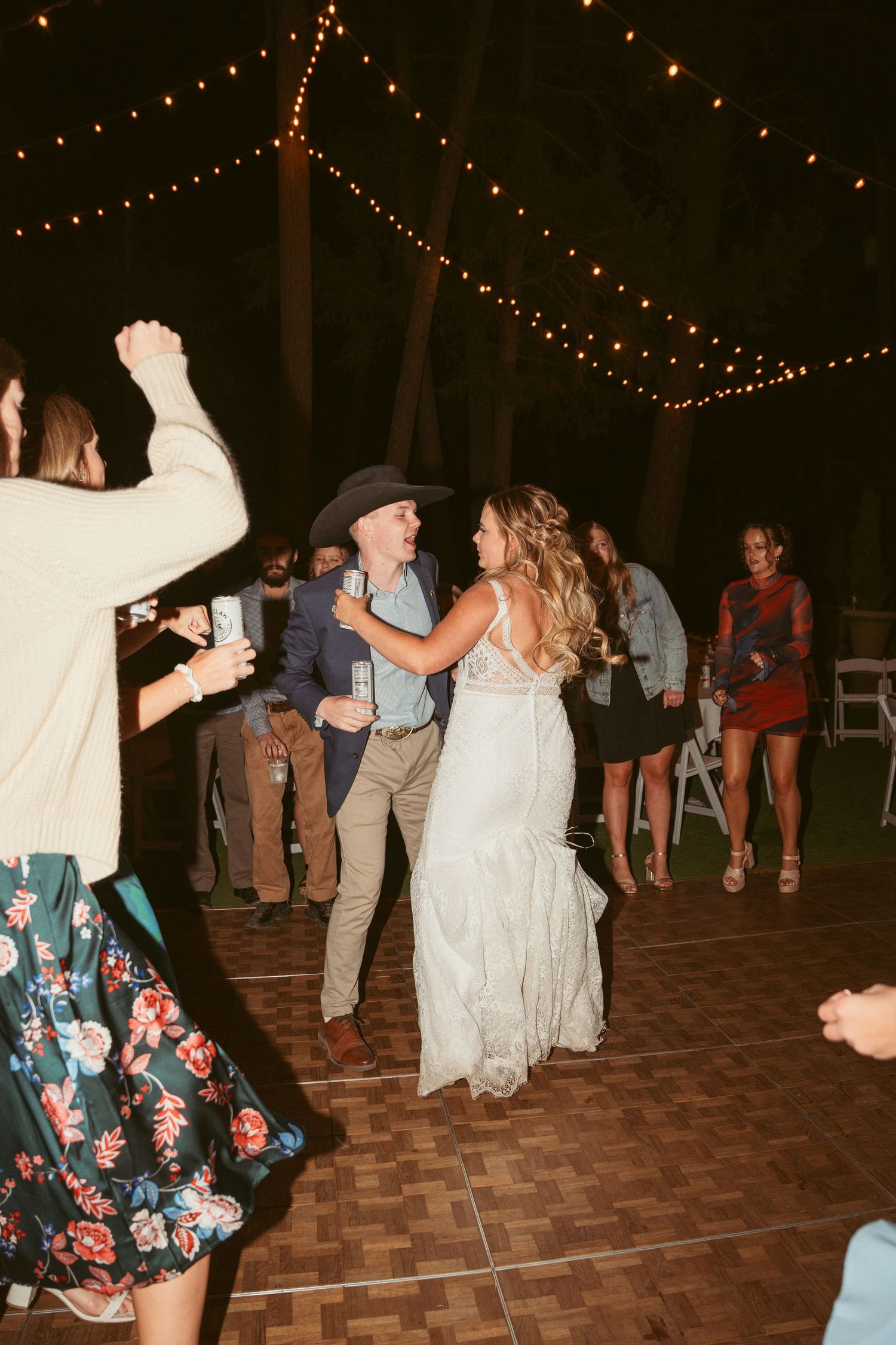 Outdoor wedding reception with guests dancing under string lights on a wooden dance floor.
