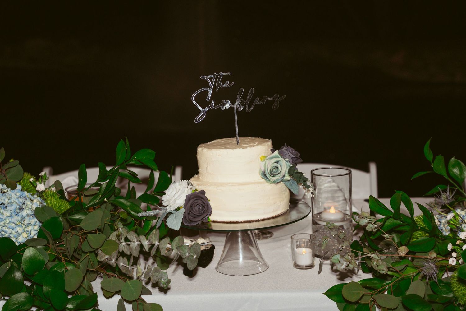 Elegant white wedding cake adorned with delicate greenery and fresh flowers sits atop a crystal cake stand.