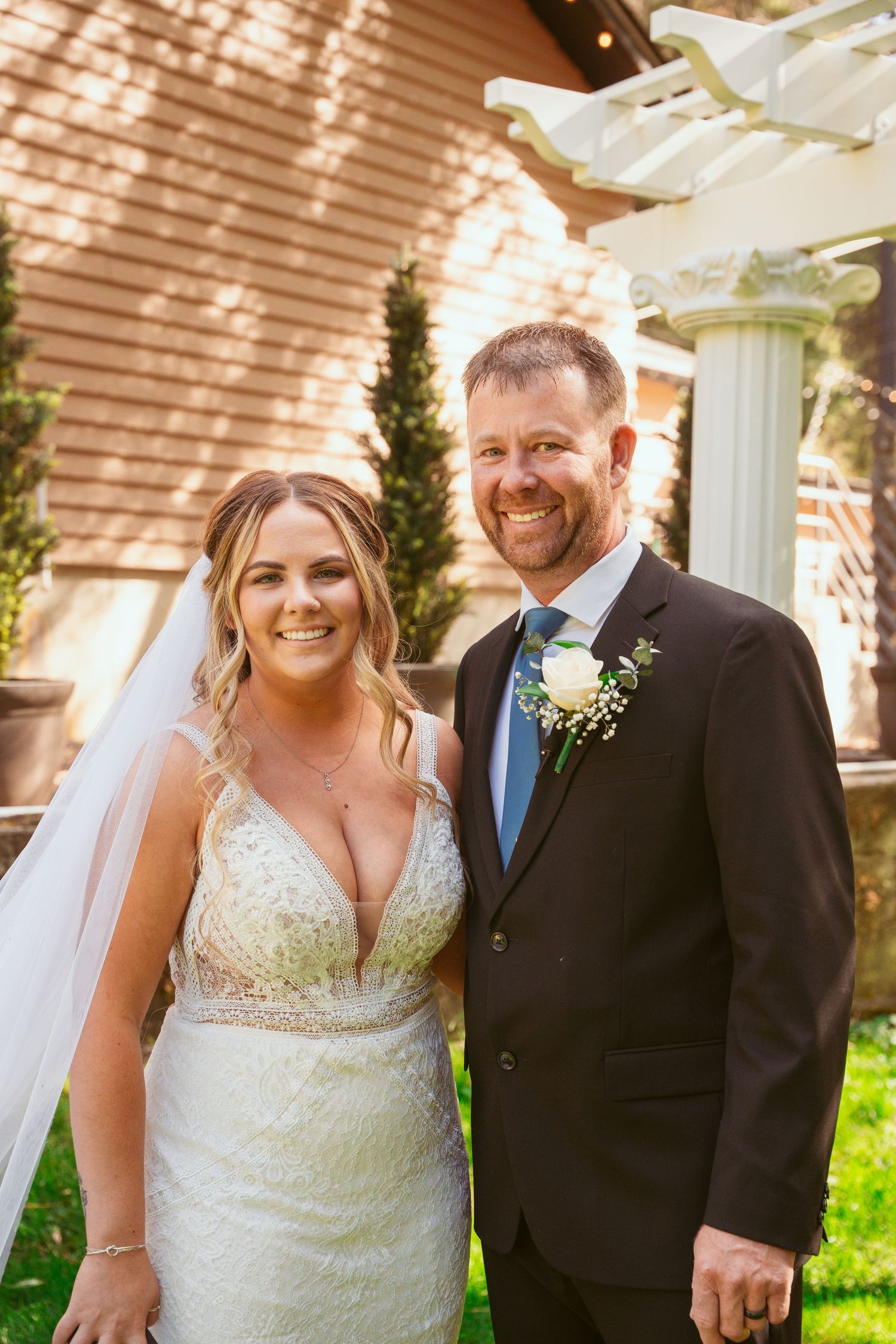 A wedding couple poses outdoors on a sunny day in front of a pergola and brick wall.