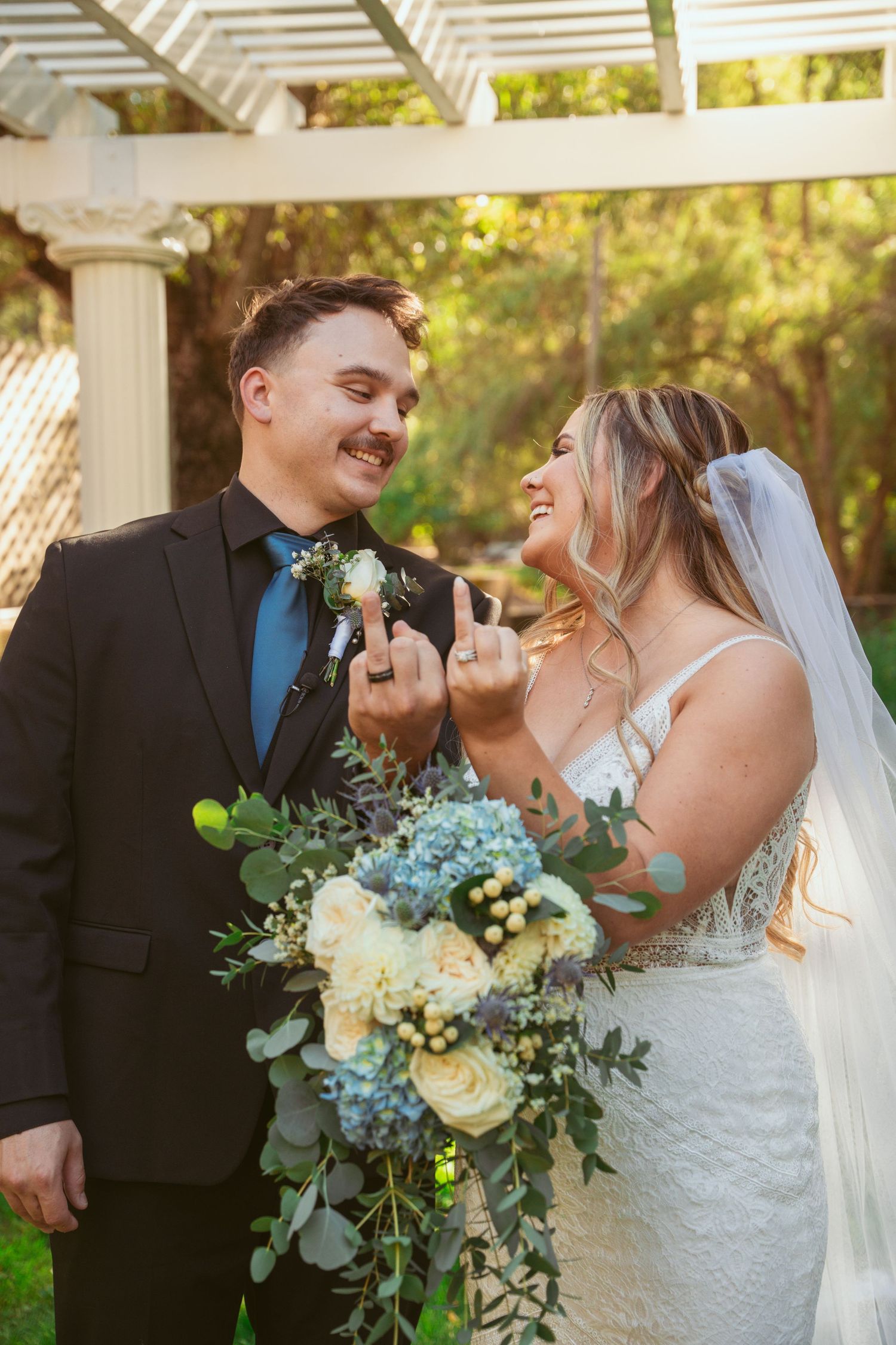 Joyful wedding couple laughing together while holding blue and white floral bouquet in outdoor garden setting.