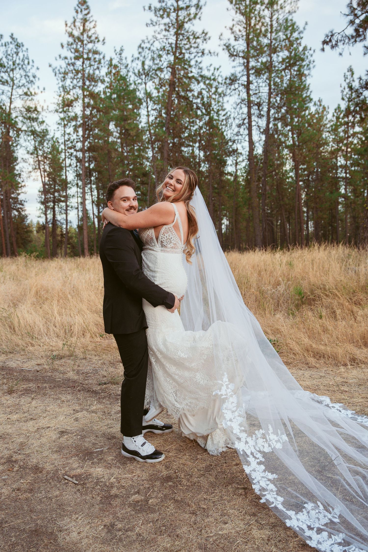 Bride's elegant lace veil trails behind as couple dances playfully on dirt path near pine forest.