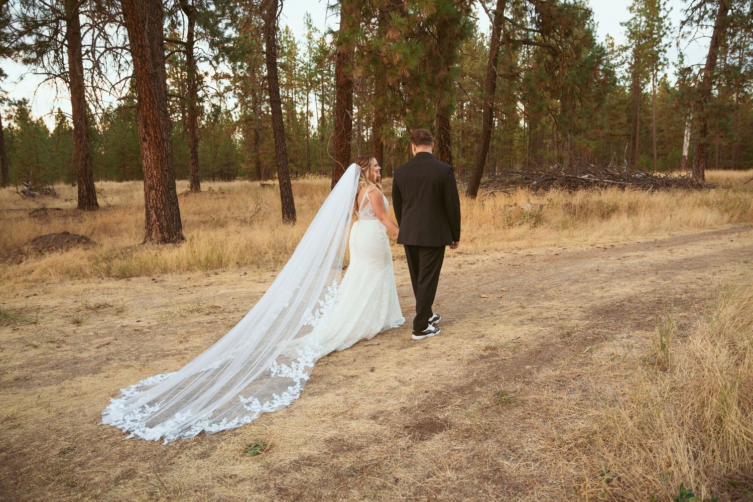 Romantic wedding couple walking through pine trees on a path with a flowing white dress train.
