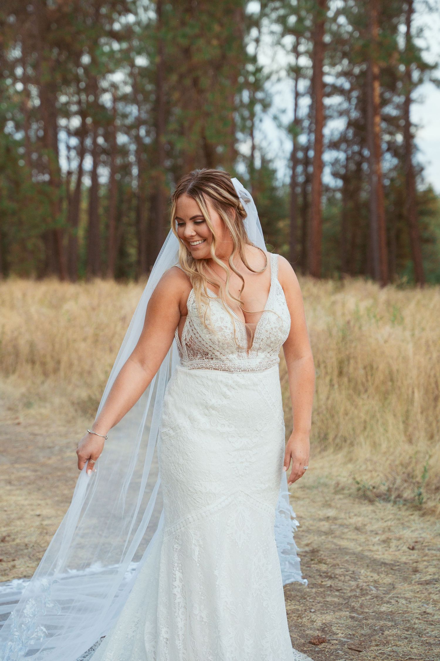 A bride in a flowing white lace dress adjusts her veil in a rustic outdoor setting with pine trees.