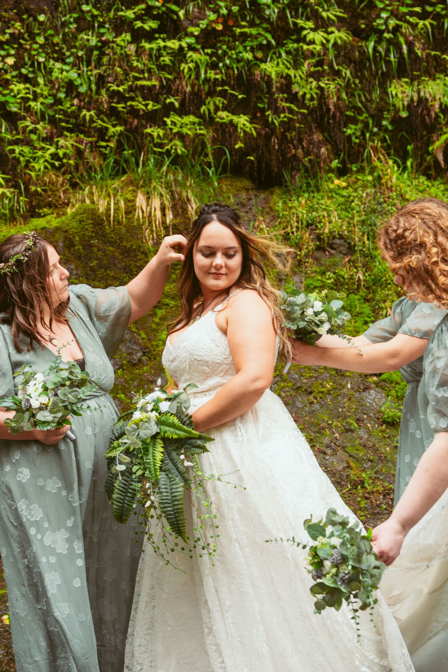 Bridesmaids in sage dresses help adjust the bride's gown by a mossy waterfall setting.