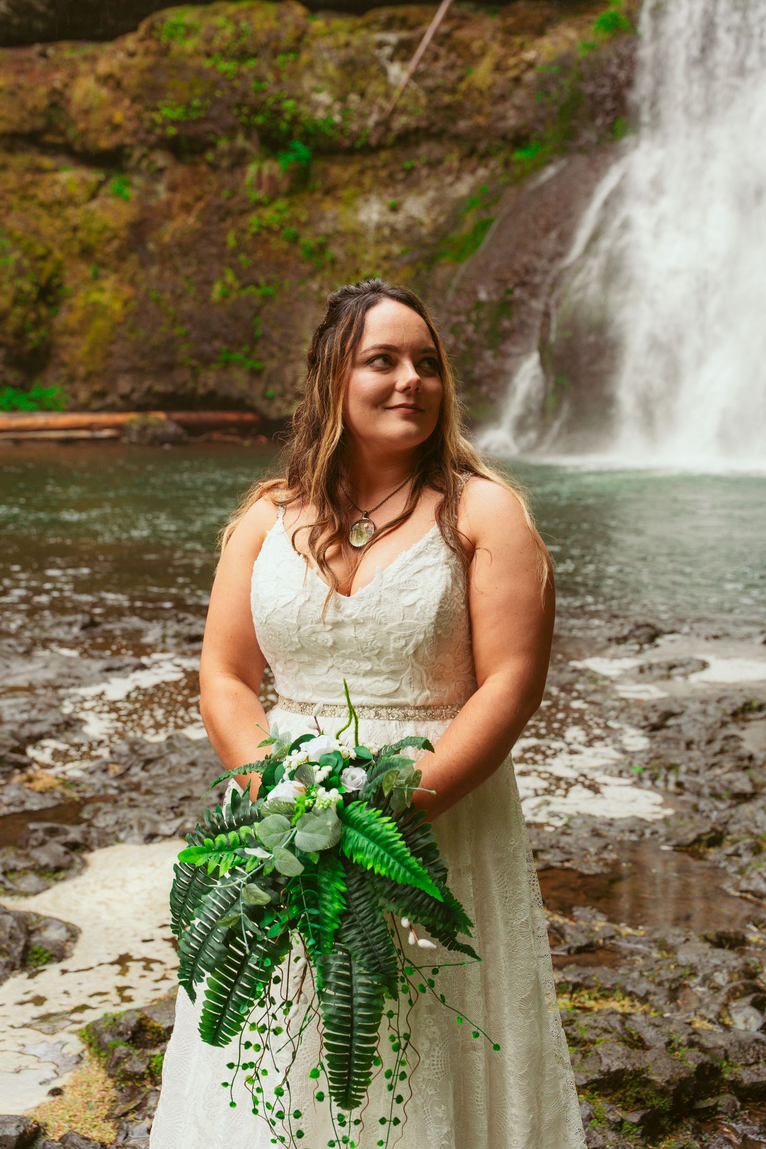Bride holds elegant bouquet of ferns and white flowers by waterfall.