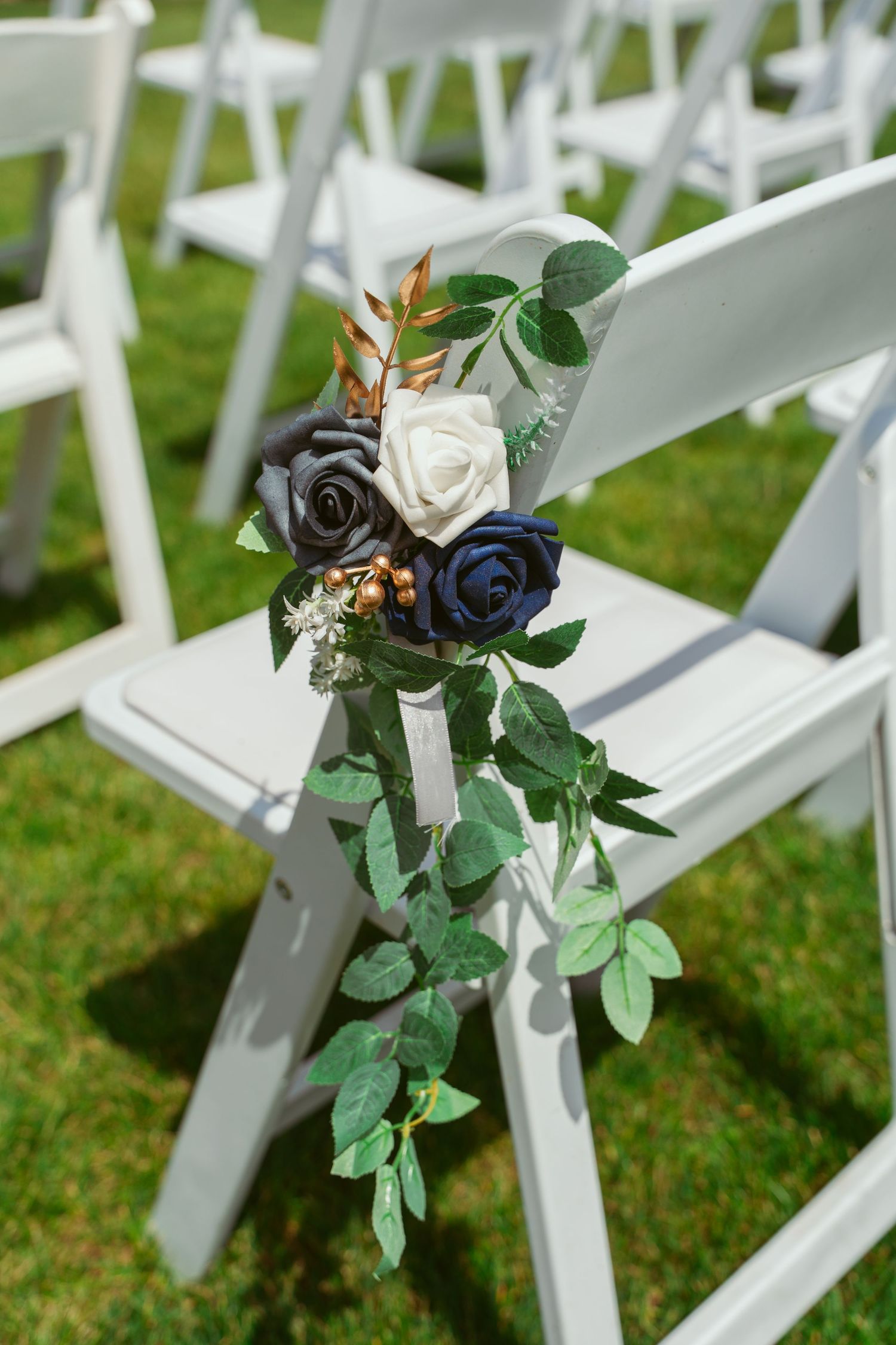 White folding chairs decorated with floral arrangements and greenery along the aisle at an outdoor wedding setup.