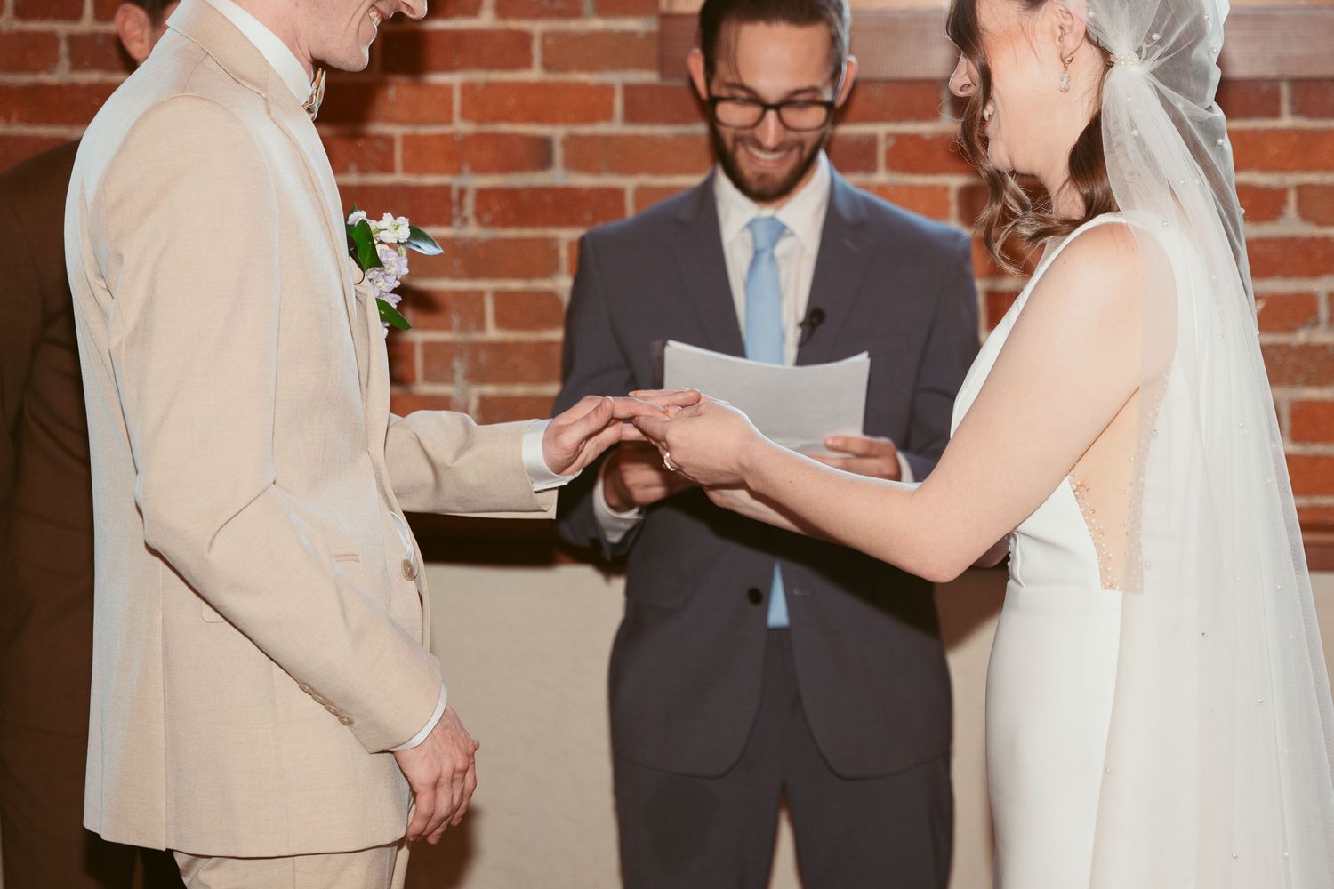 Wedding officiant leads ceremony while couple stands before guests in brick-walled venue.