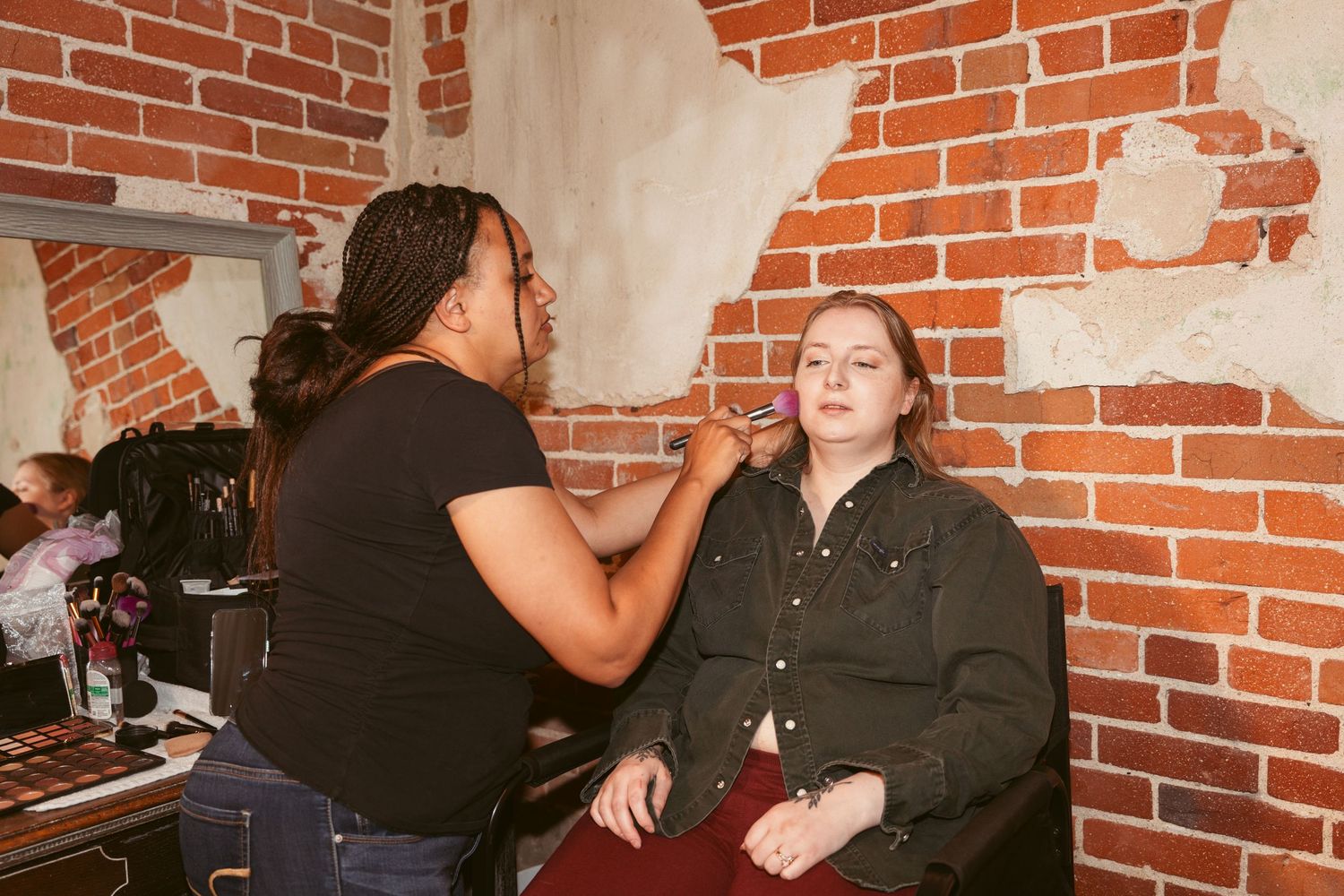 Professional makeup application session in progress against rustic brick backdrop.
