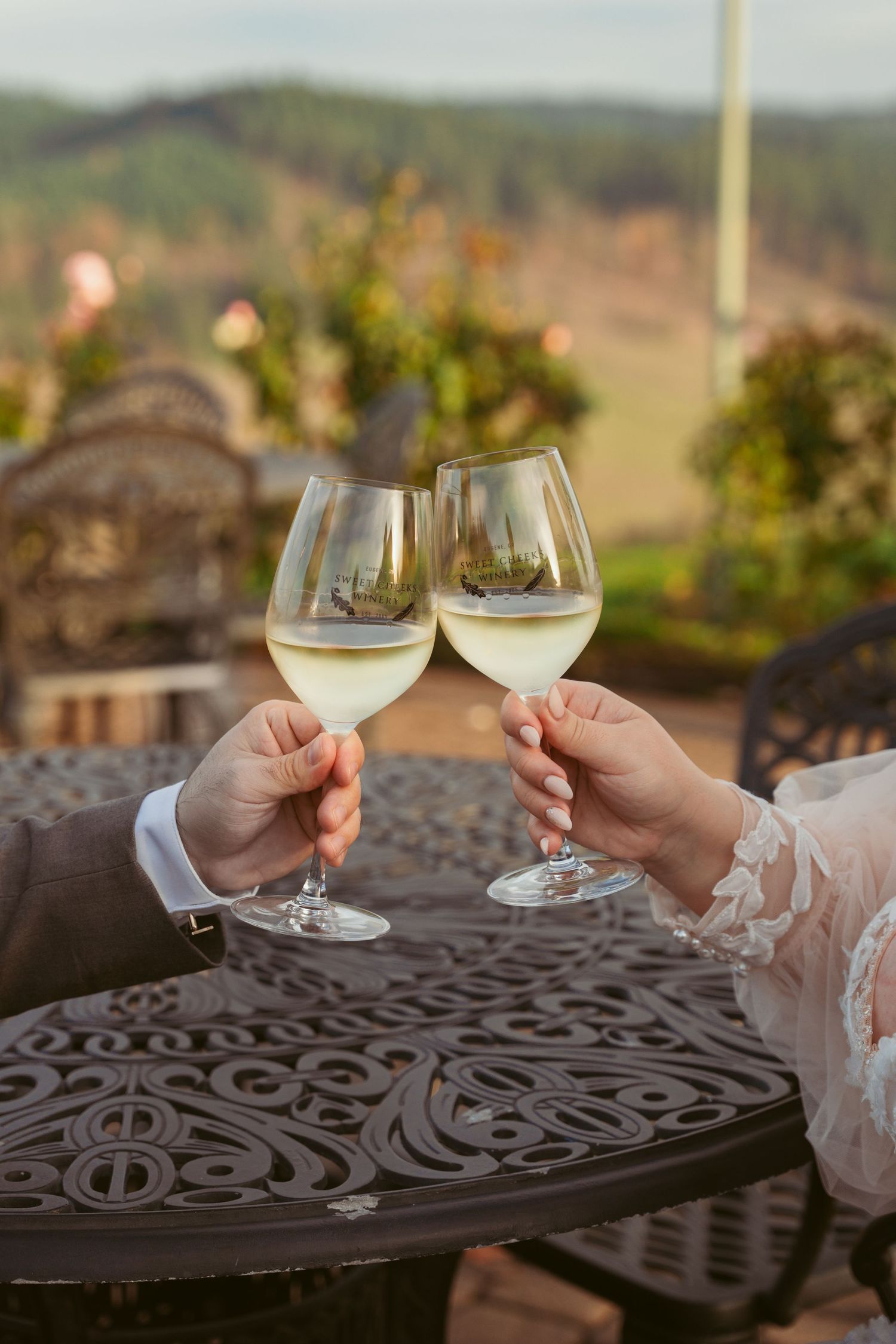 Two wine glasses clink together against ornate metal table with vineyard backdrop.