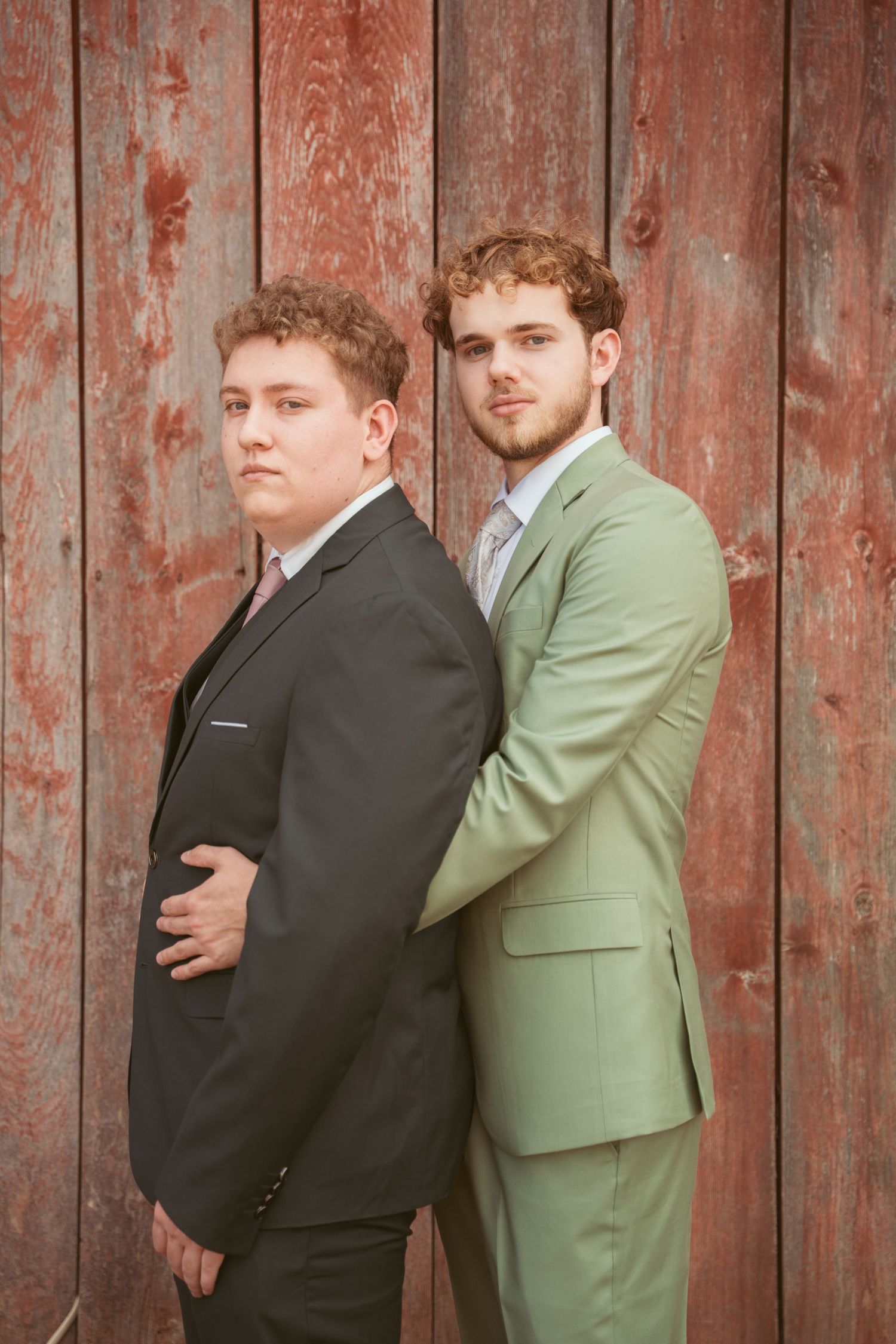 Two men in contrasting suits stand close together against weathered red barn siding.