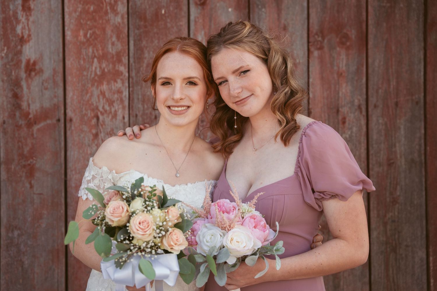 Bridal party in dusty rose dresses gather for photos against weathered wooden barn wall.