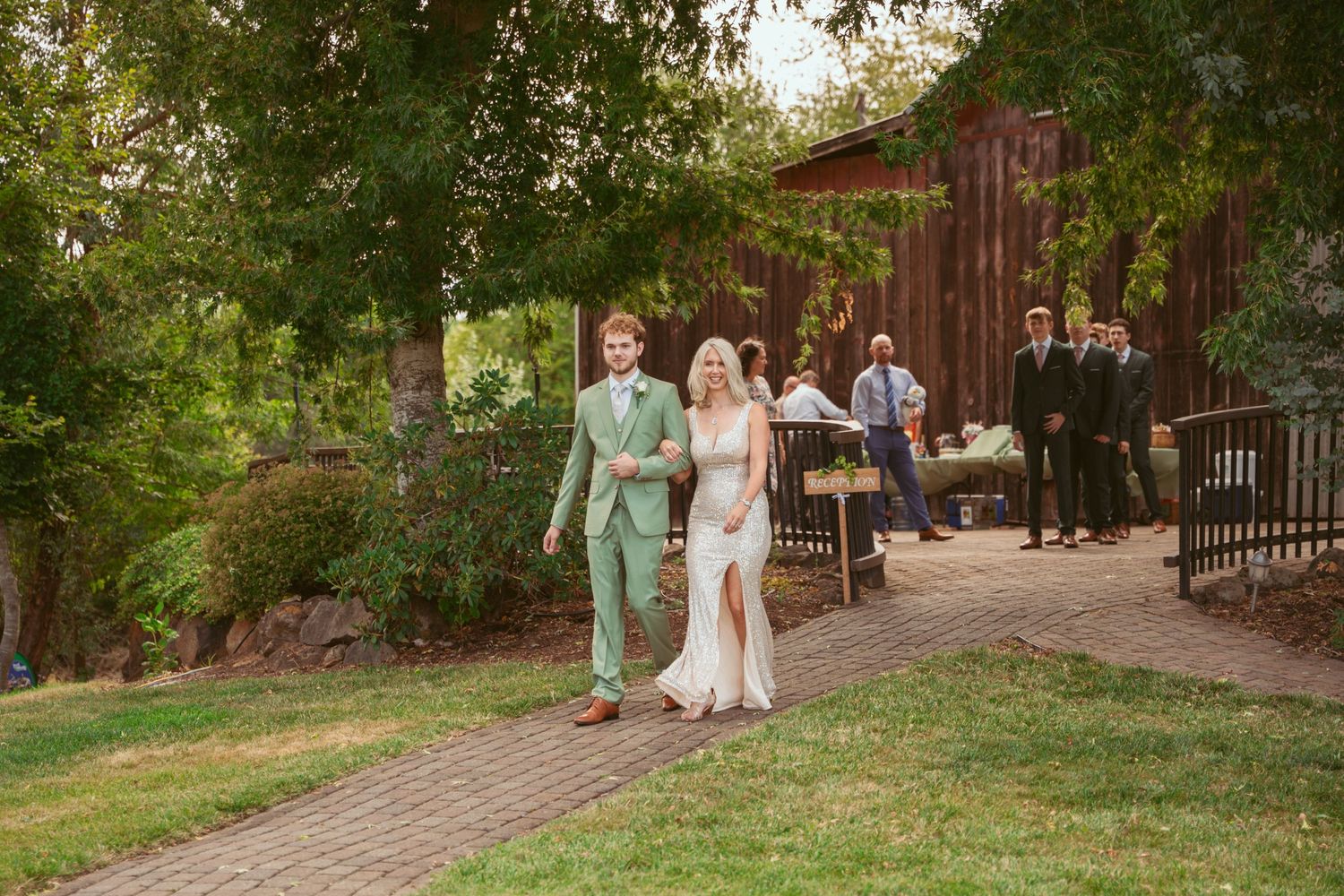 Wedding party walks along a brick path outside a rustic red barn venue on a sunny afternoon.