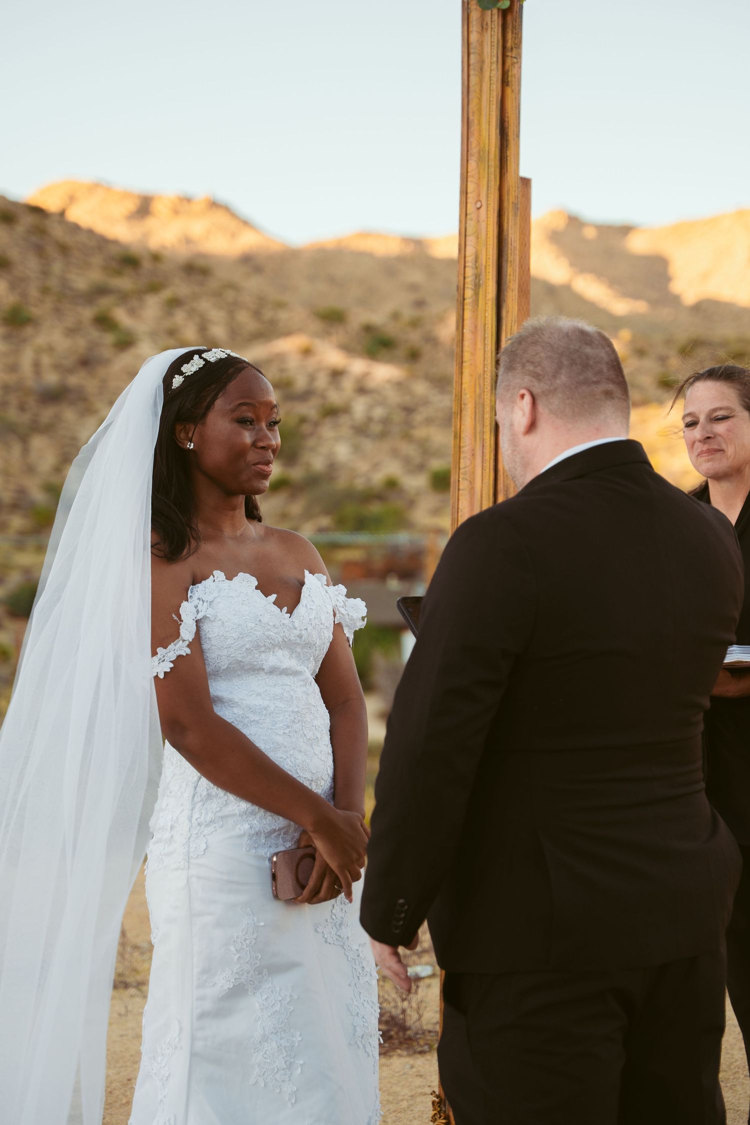 Wedding ceremony in progress under wooden arch with desert mountains in background.