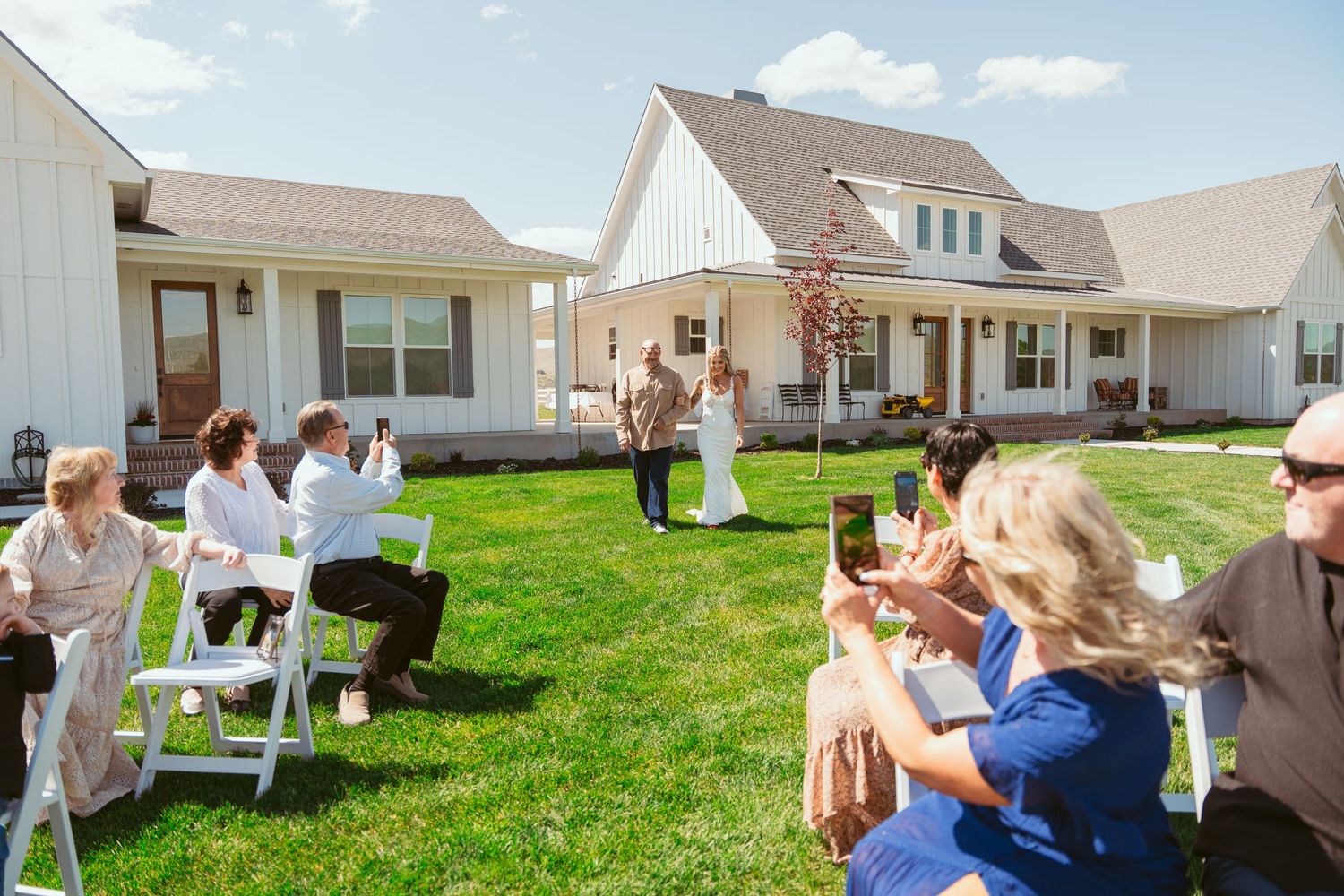 Outdoor wedding ceremony setup with white chairs on a green lawn against a backdrop of mountains and blue sky.
