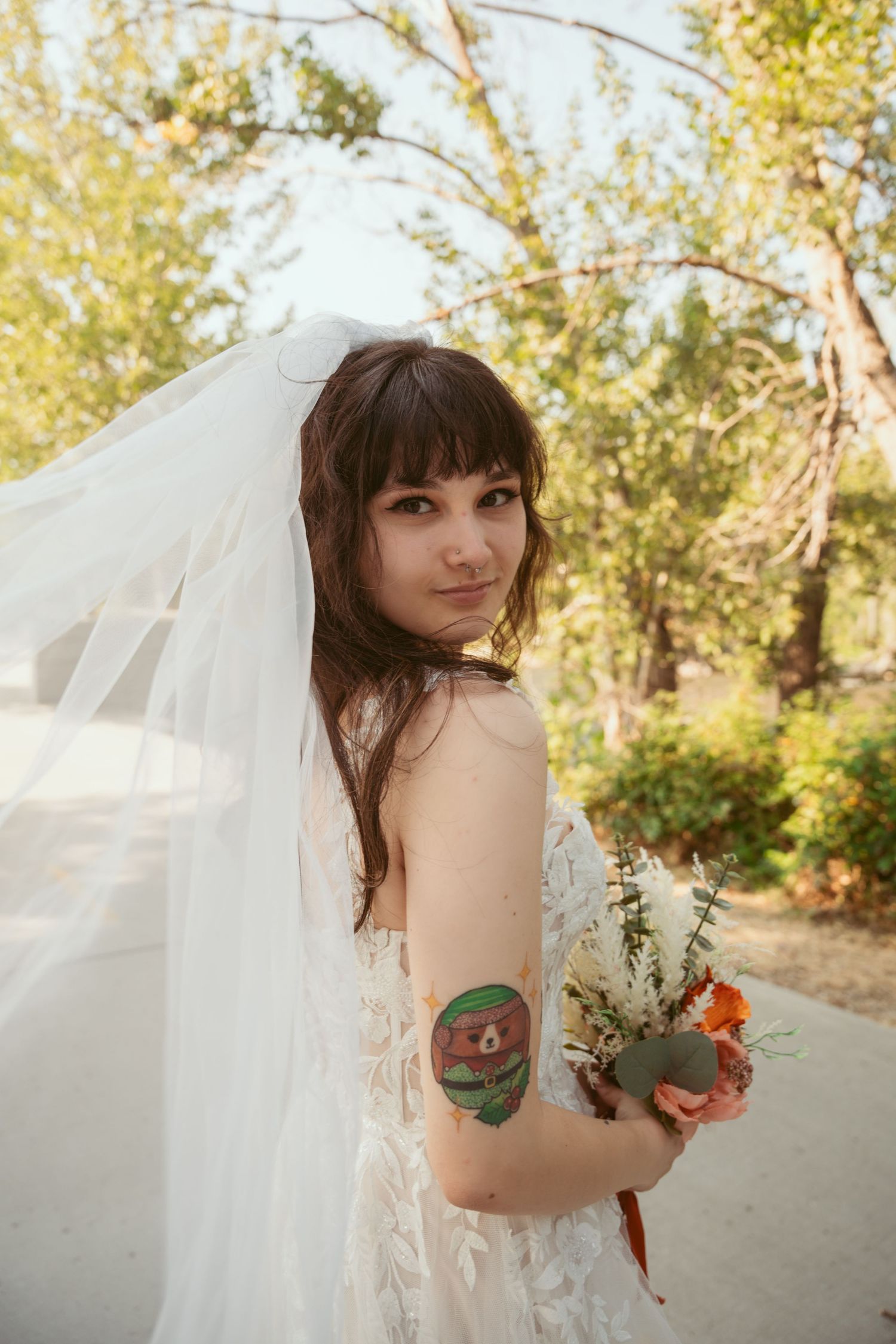 A bride in a flowing white dress poses with flowers against a sunlit natural backdrop.