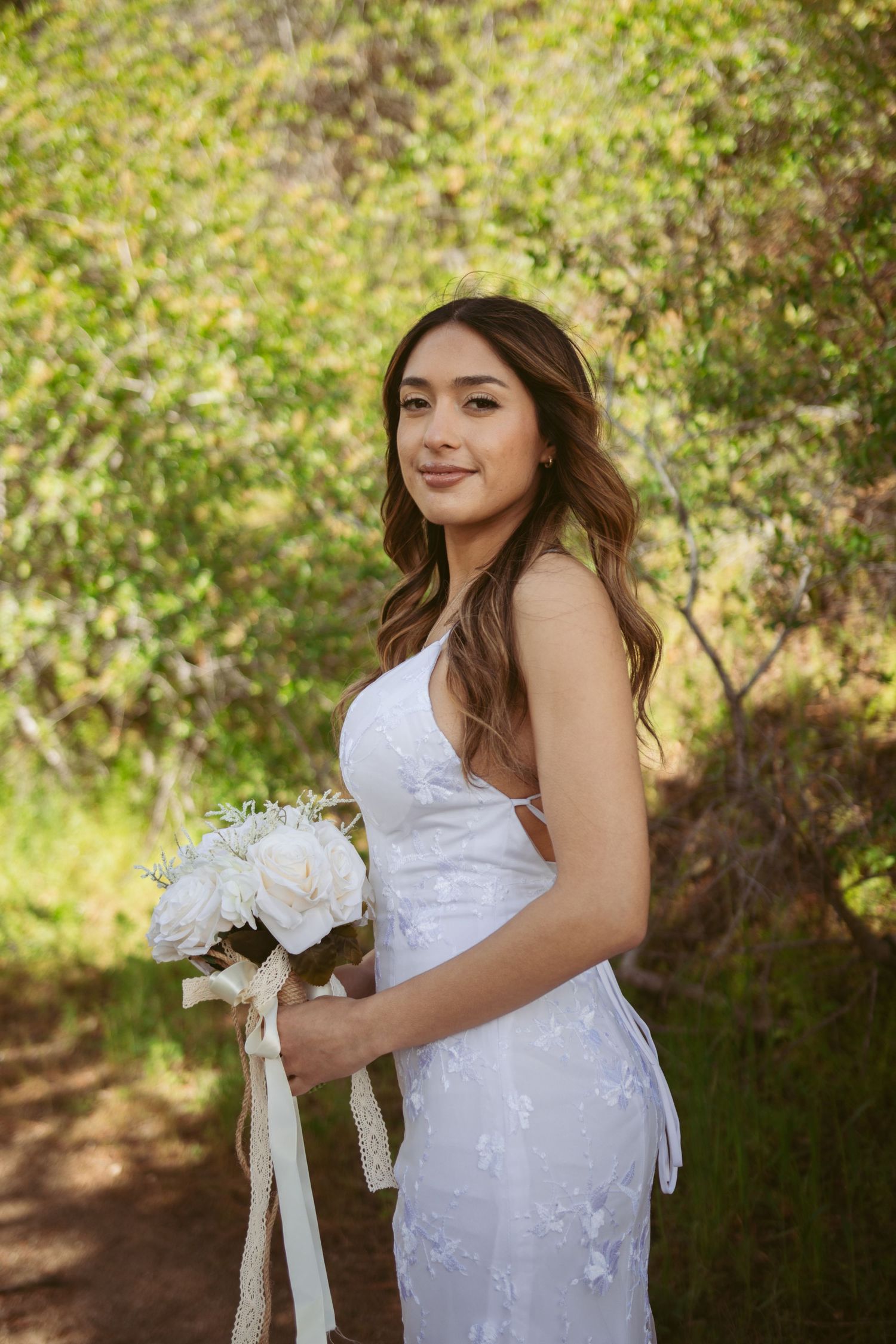 A bride in a white dress holds a bouquet while standing in front of lush greenery.