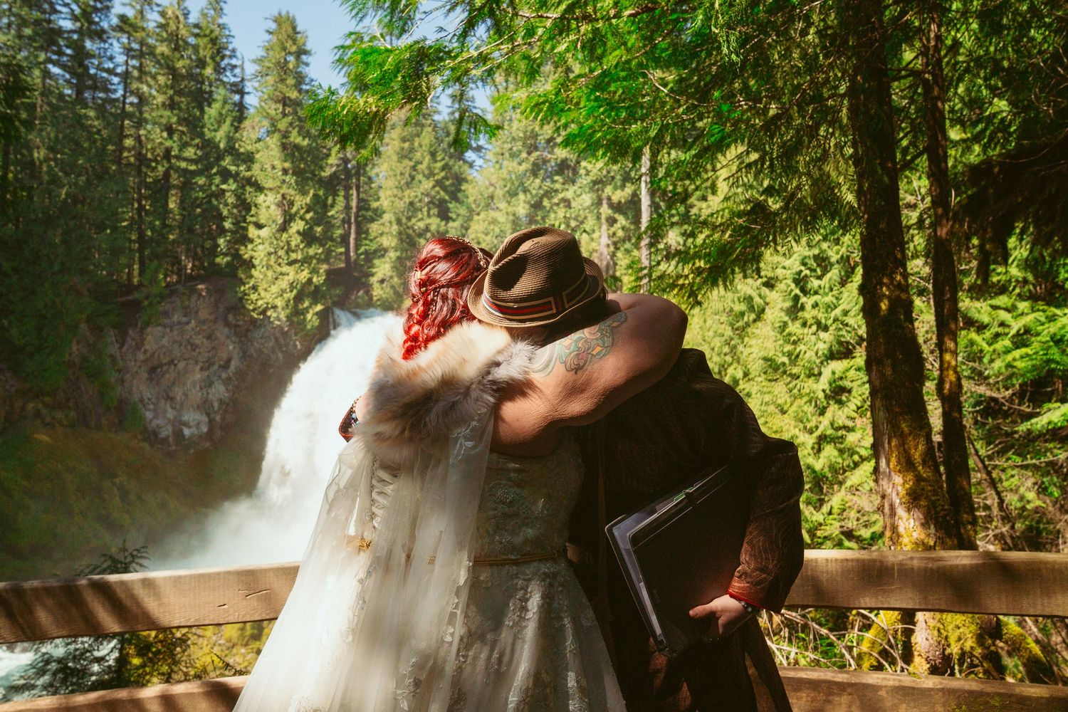 A wedding couple embraces near a majestic waterfall in a lush forest setting.