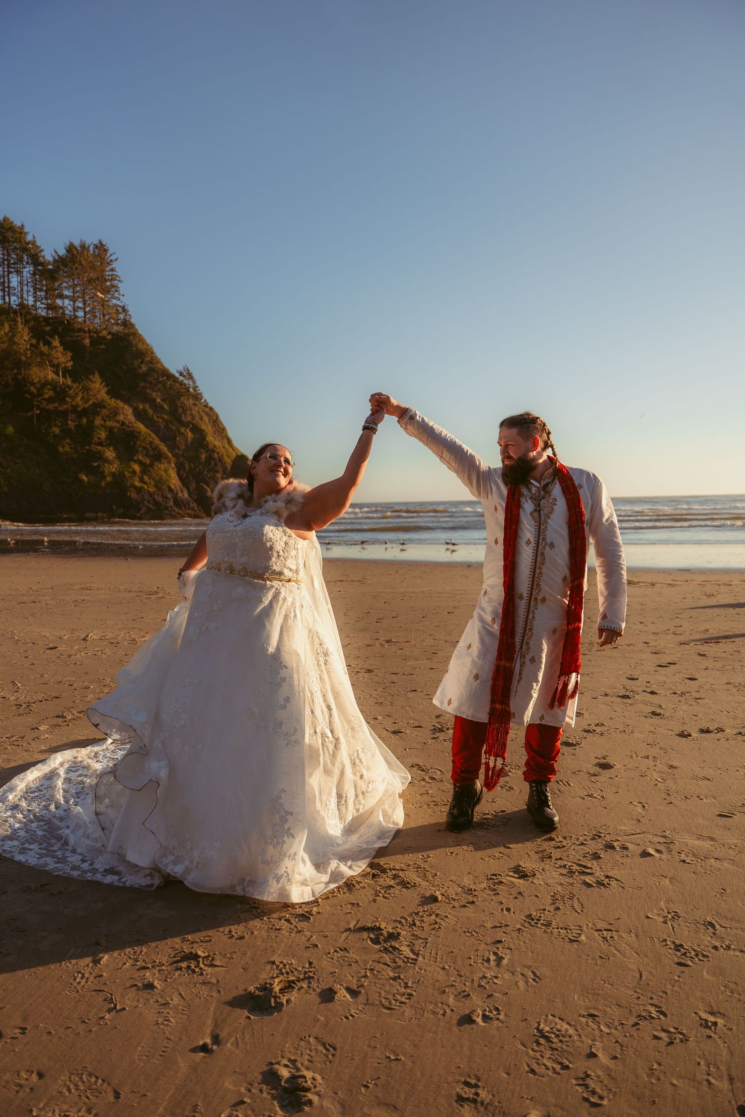 A couple in wedding attire dances joyfully on a beach with dramatic cliffs and ocean waves behind them.