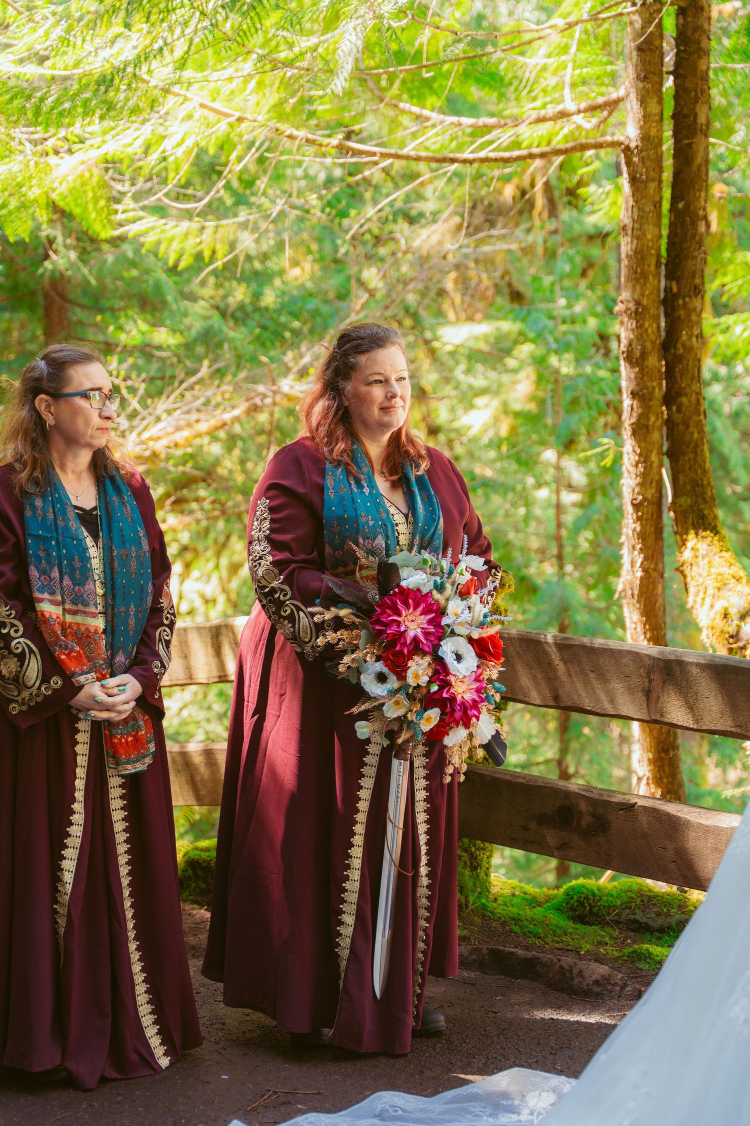 Bridesmaids in burgundy medieval-style dresses hold bouquets during an outdoor forest ceremony.