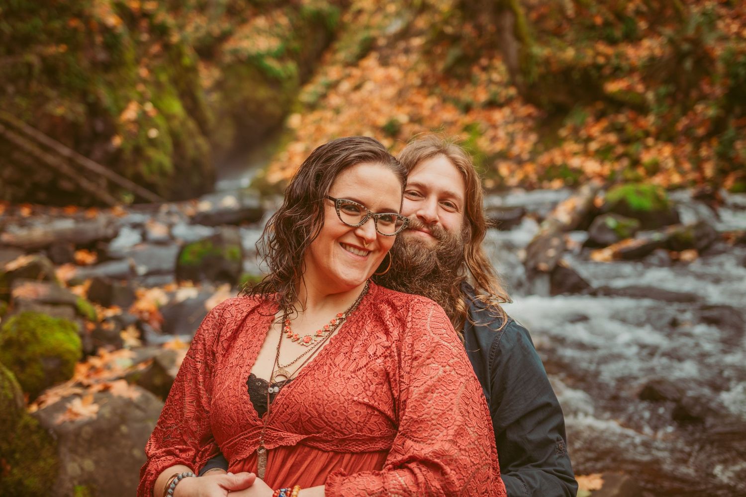 A couple in autumn attire shares a tender moment by a flowing stream.