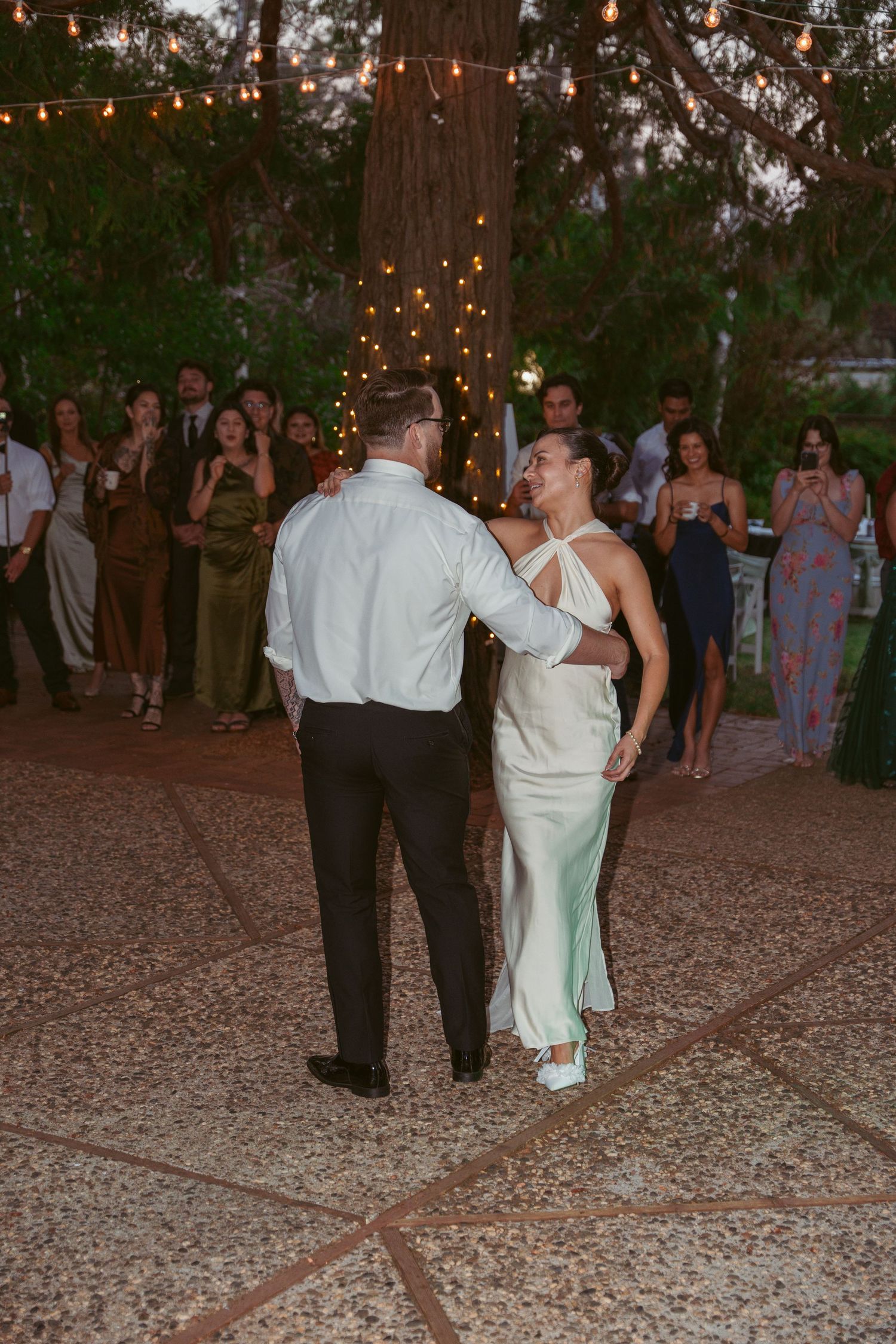 Wedding guests dance together on an outdoor patio decorated with string lights at dusk.