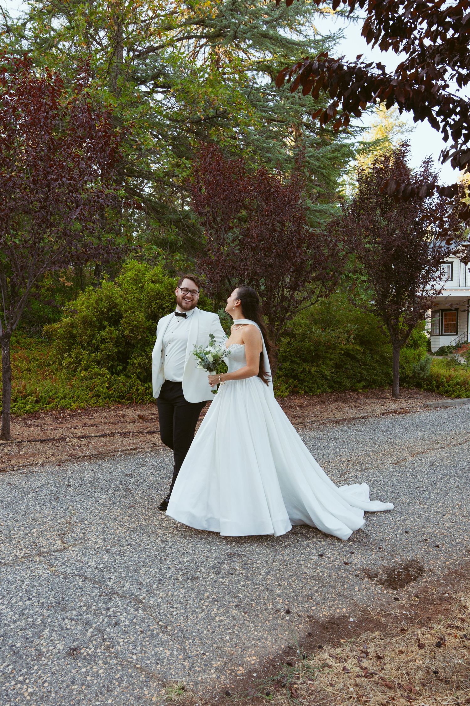 A couple in wedding attire poses along a gravel path surrounded by lush greenery.