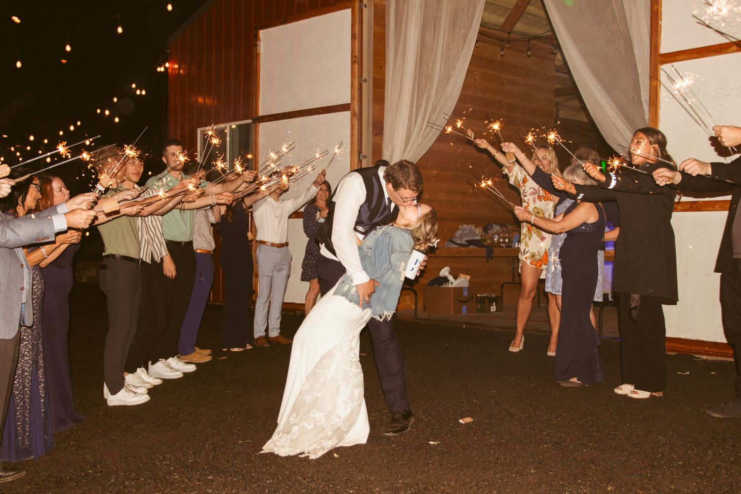 Newlyweds share romantic dip kiss during sparkler exit at wedding reception with guests forming tunnel.