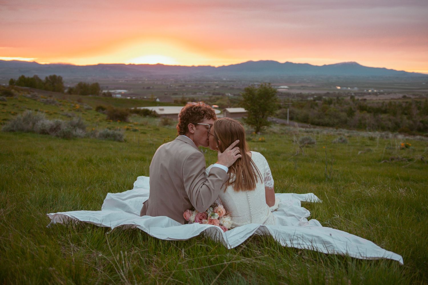 A couple shares an intimate moment on a blanket during sunset in a scenic mountain field.