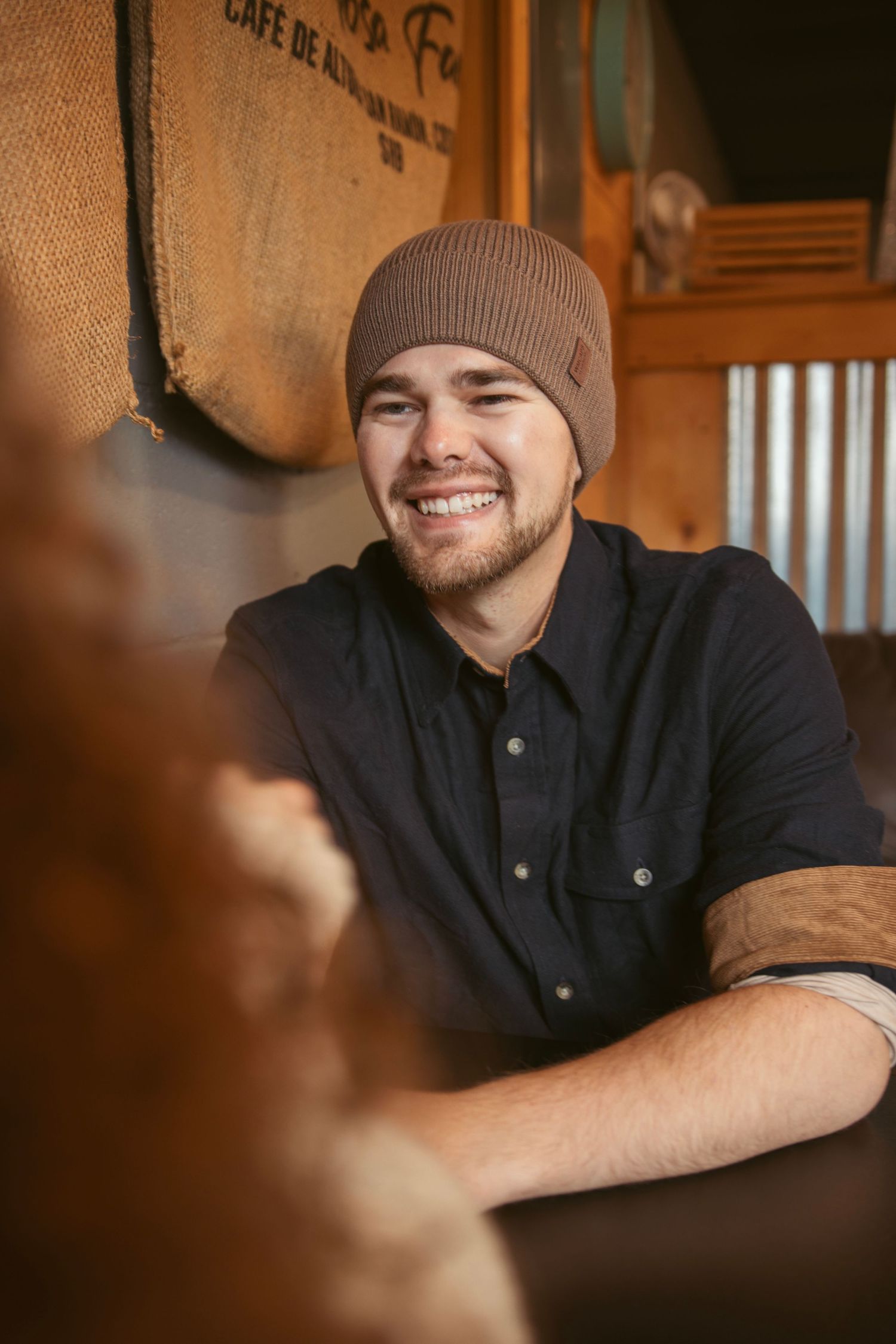 Person in beanie and black jacket smiling in warm cafe lighting.