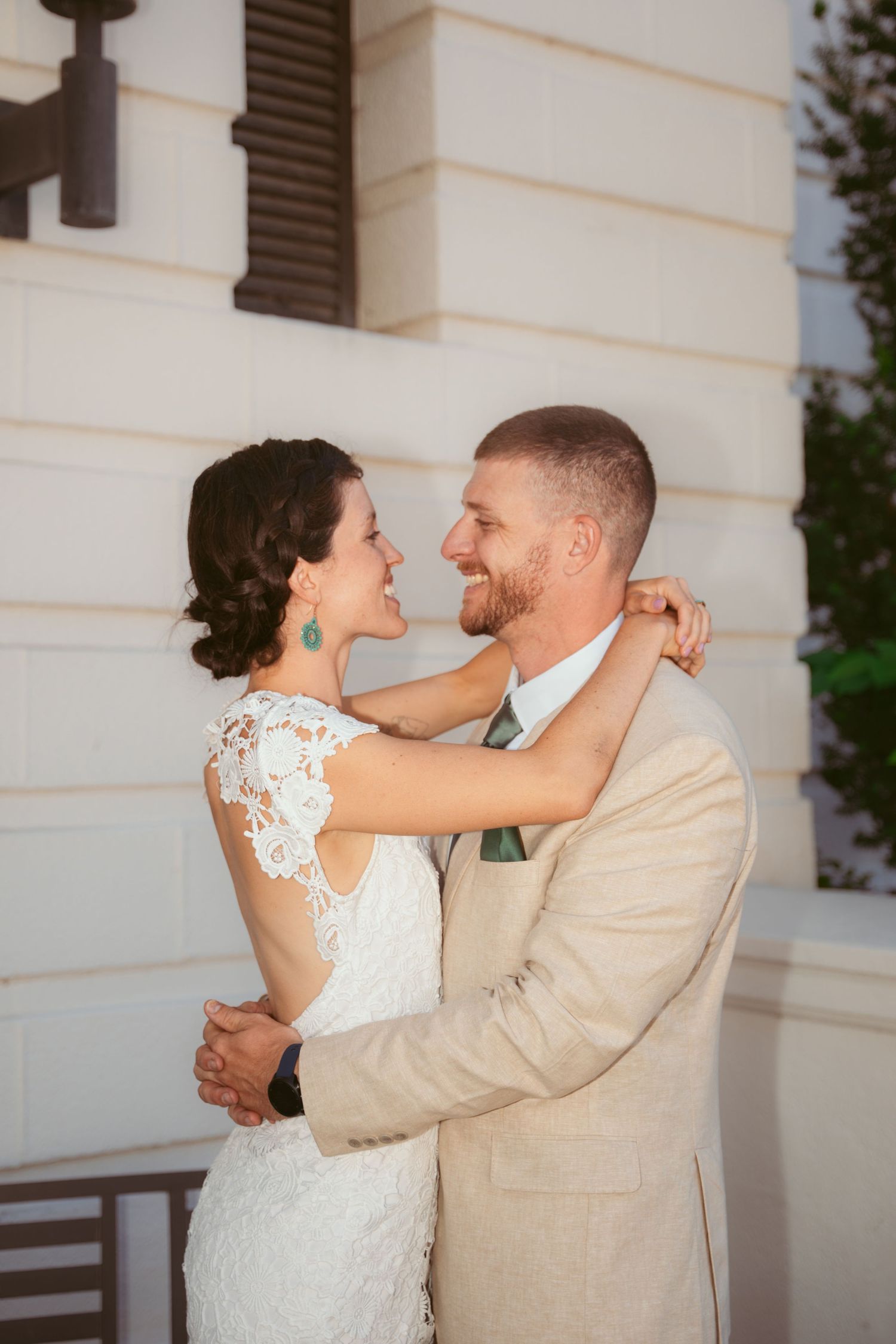 Tender moment between newlyweds in tan suit and lace dress against white wall.