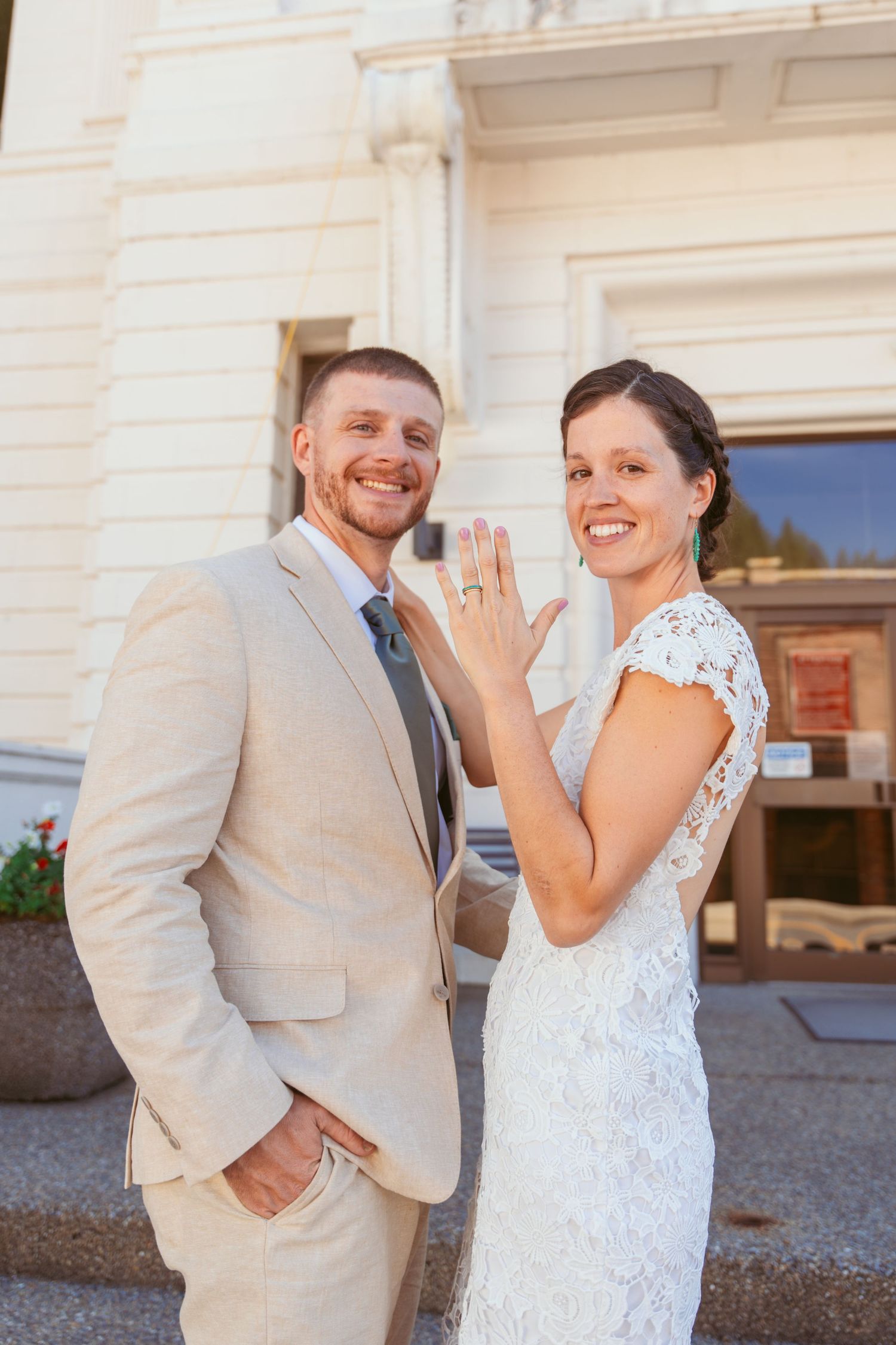 Happy couple in beige and white attire showing off engagement ring outside building.