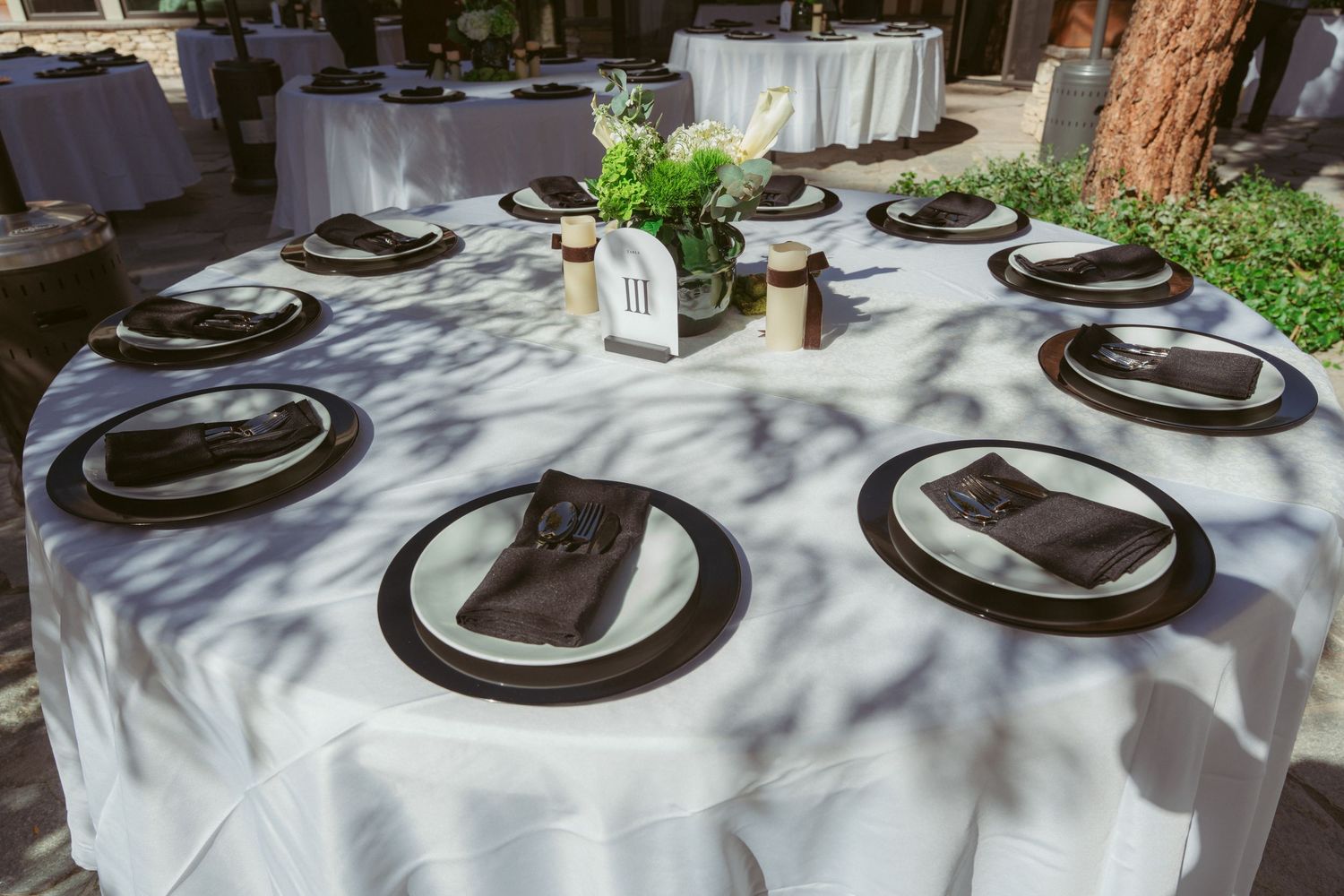 Elegant table settings with white linens and dark charger plates in an outdoor setting.