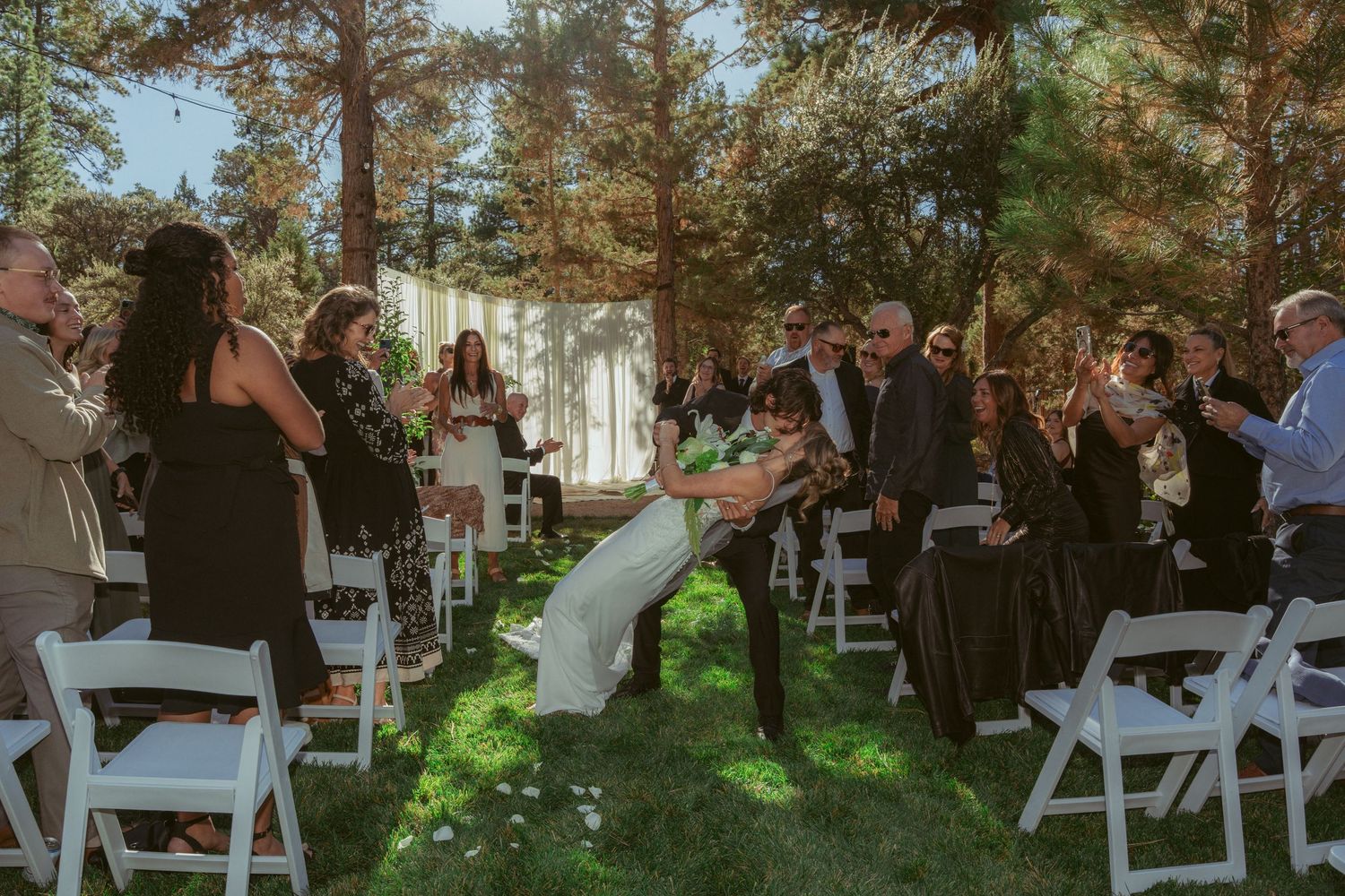 Outdoor wedding ceremony with guests seated on white chairs under tall pine trees.