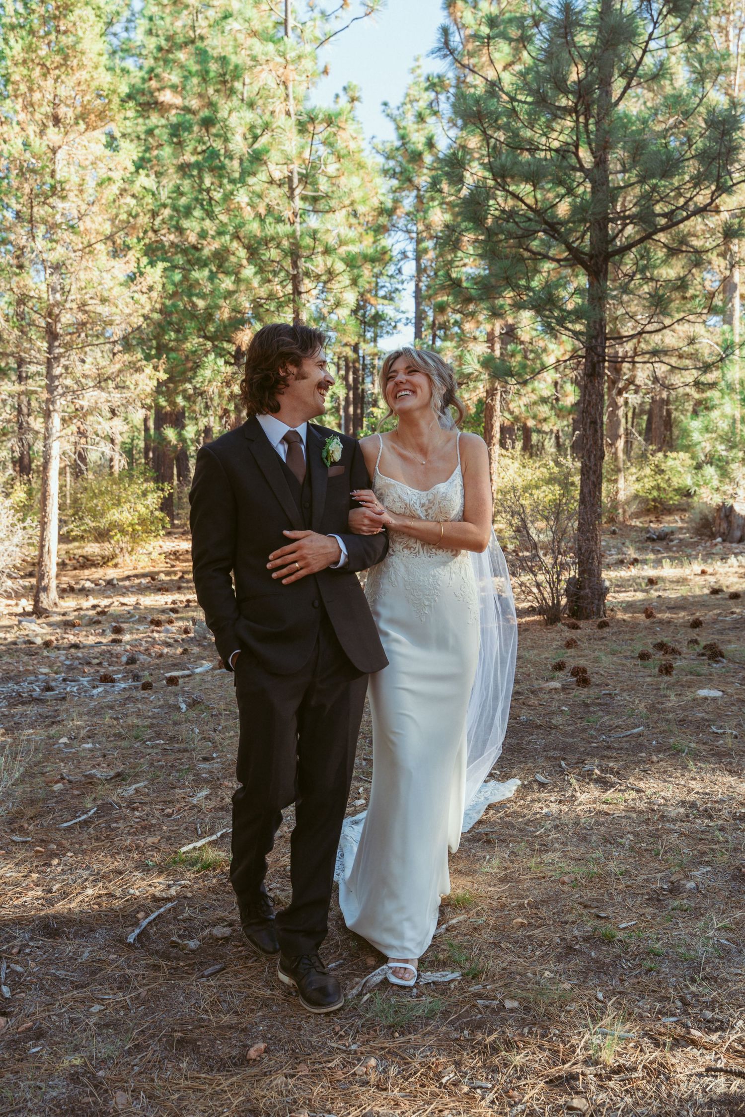 A playful wedding couple dances together among tall pine trees in a forest setting.