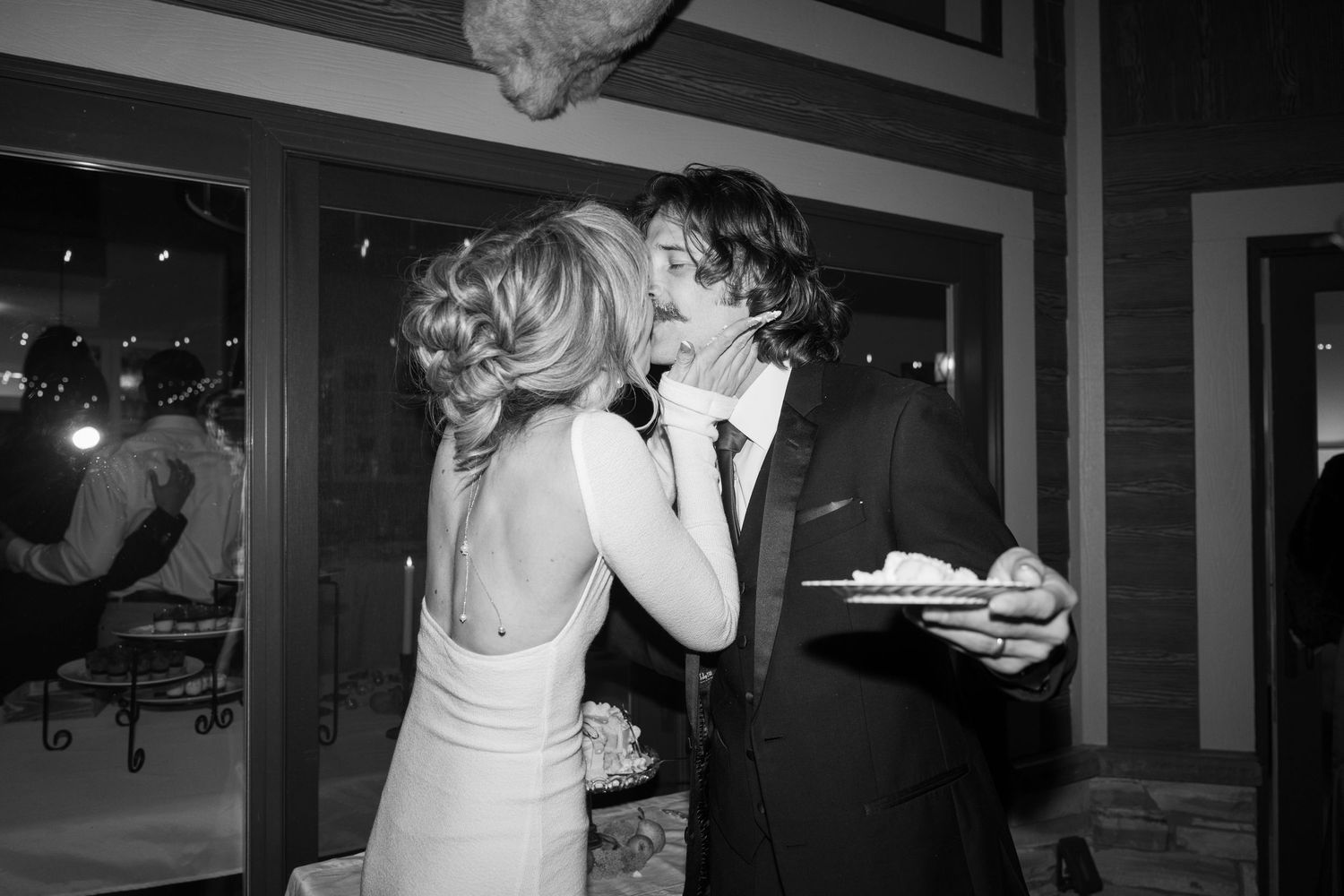 Bride in white dress and groom in dark suit share wedding cake at evening reception.