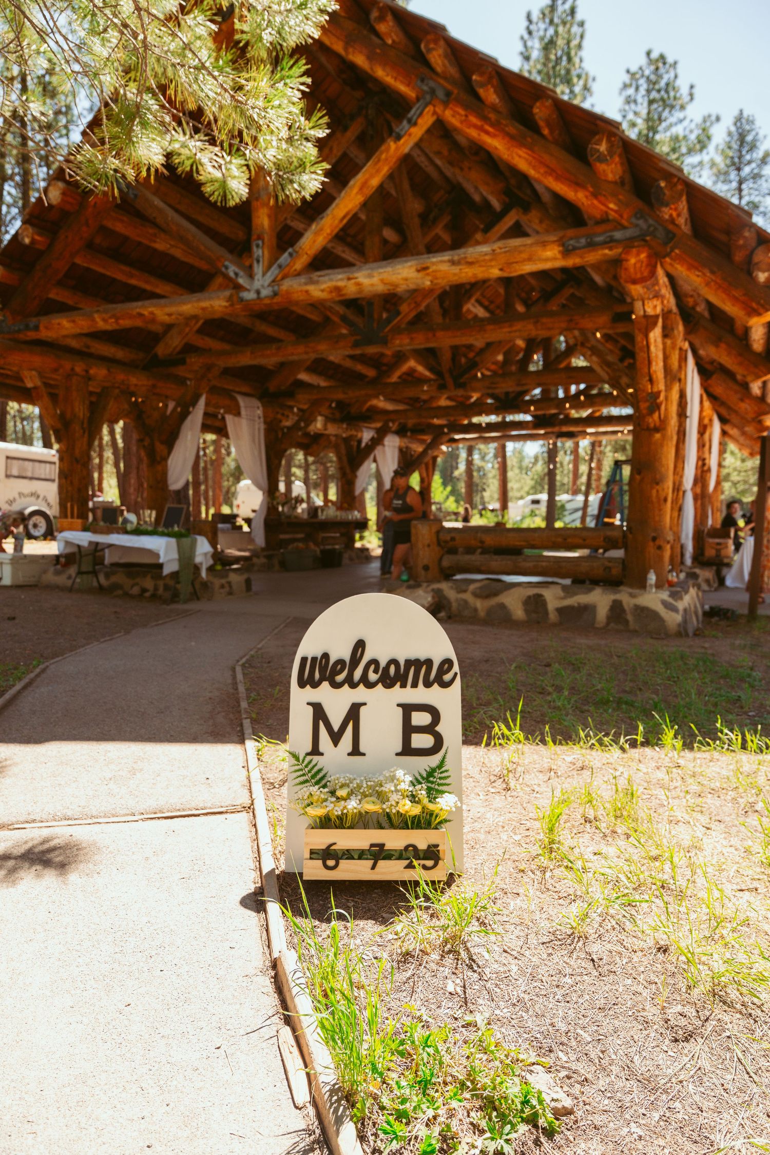 Rustic wooden pavilion entrance with a welcome sign and natural log beam construction in an outdoor setting.