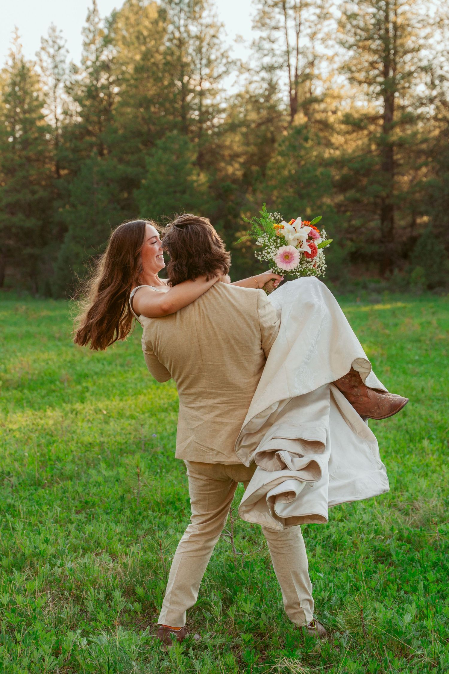 A couple spins joyfully together in a green field with evergreen trees in the background.