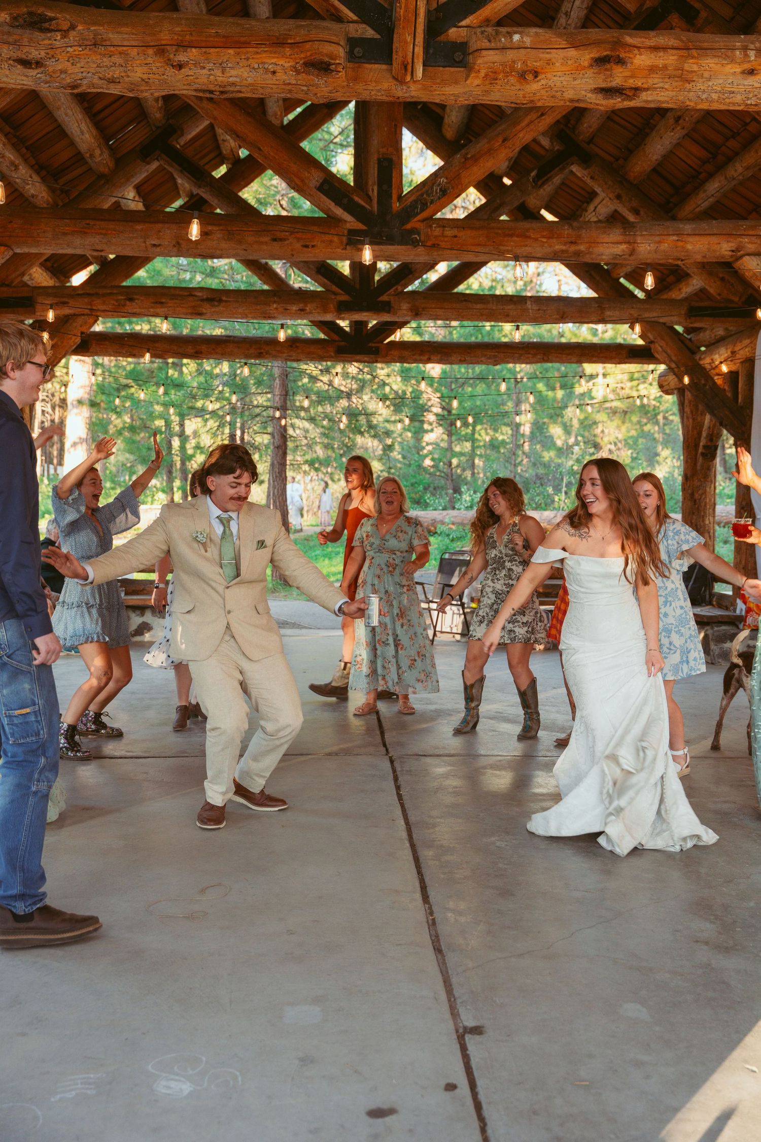 Large group dancing at an outdoor wedding reception under a wooden shelter.