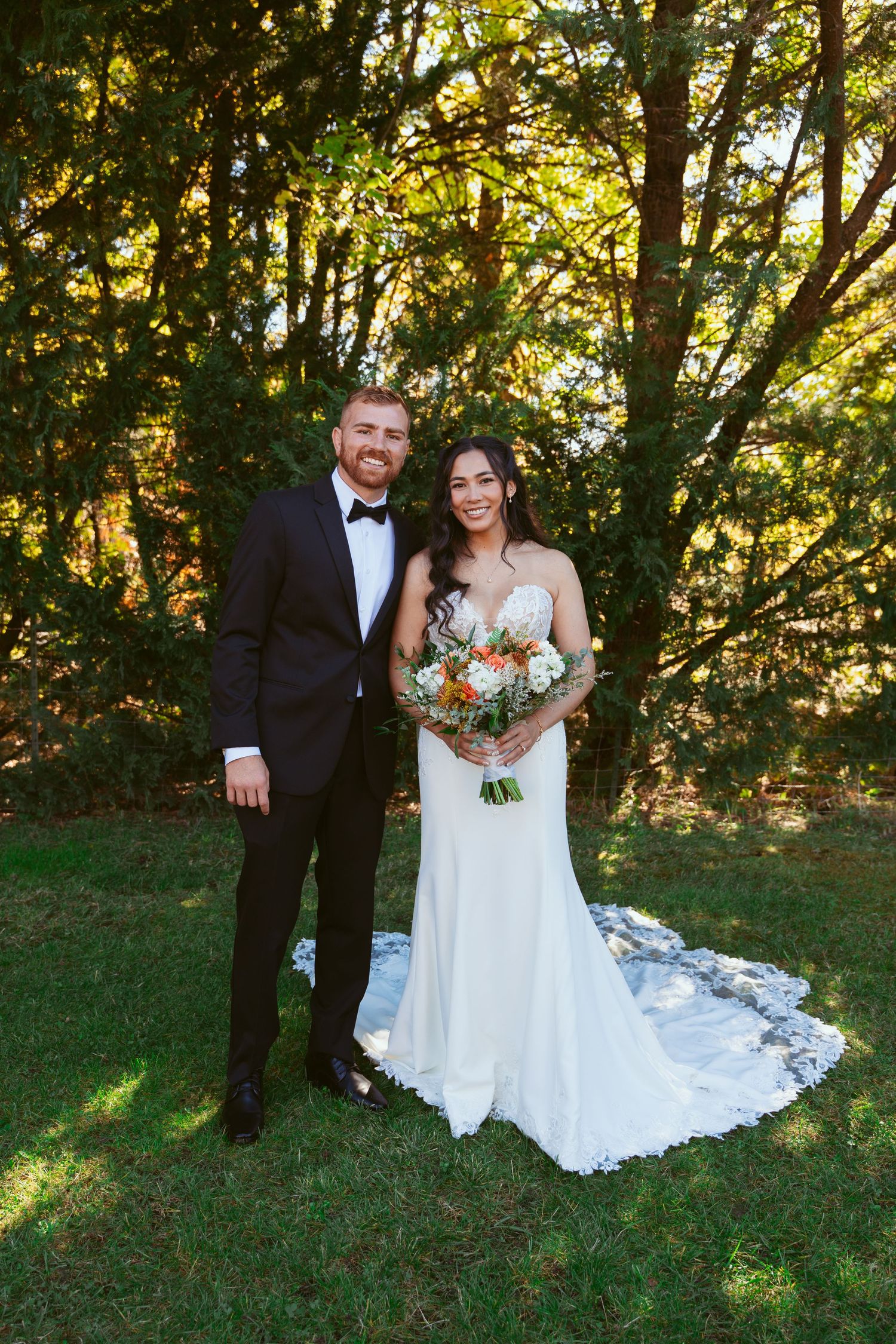Wedding portrait of newlyweds standing together on lawn with autumn trees and golden sunlight.