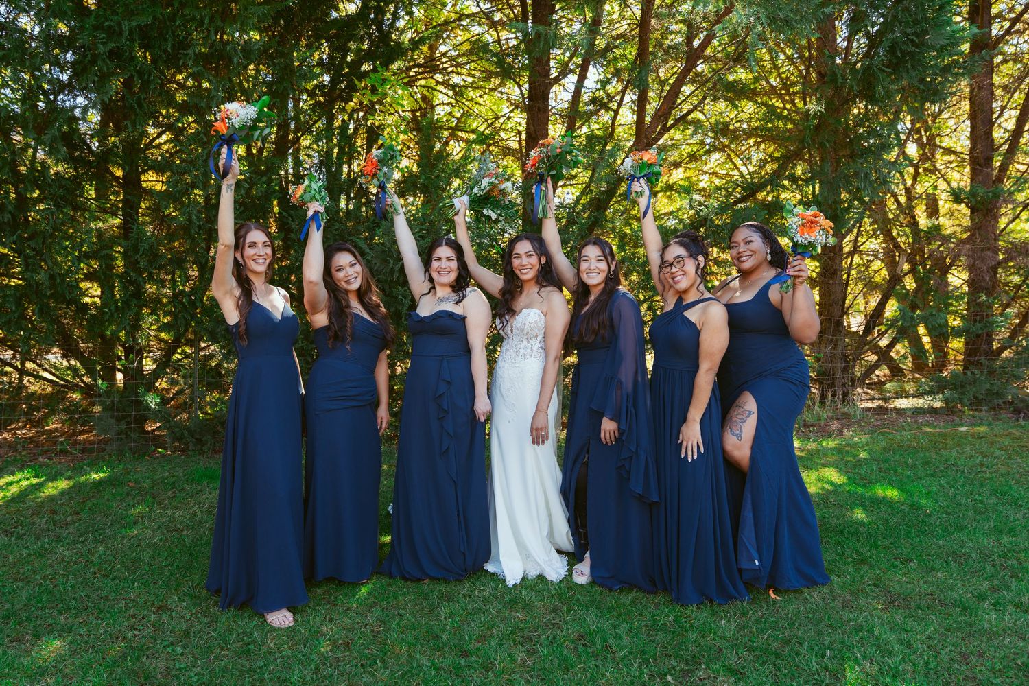 Wedding party in navy blue dresses poses joyfully outdoors with bouquets raised in celebration.