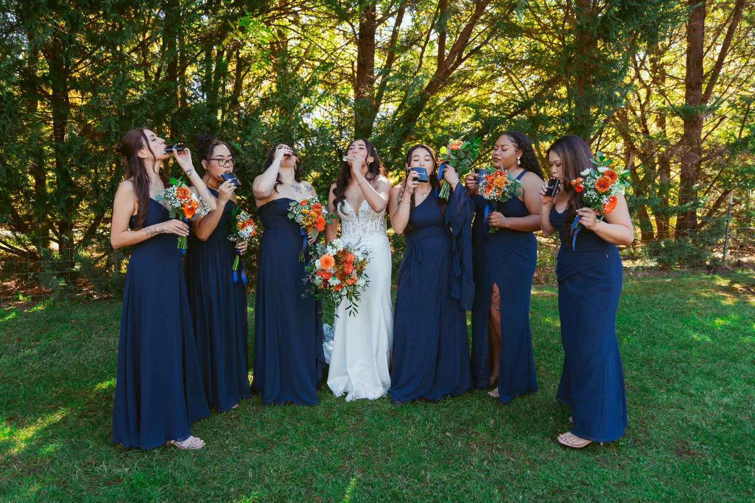 Wedding party in navy dresses shares laughs while holding bouquets in wooded setting.