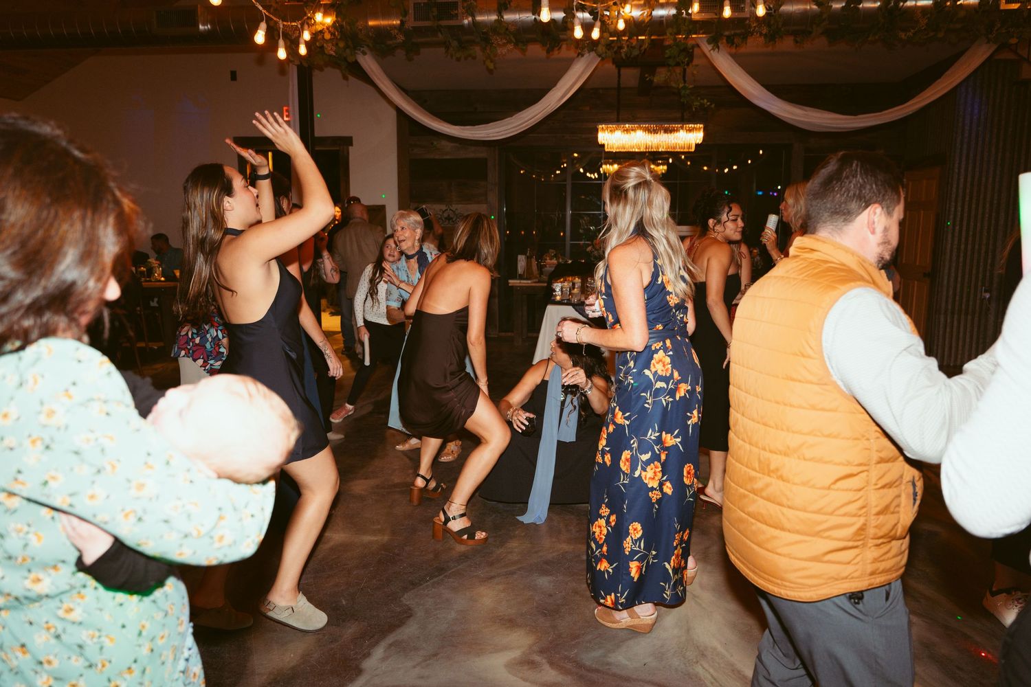 Wedding guests dancing and celebrating at a reception with string lights and decorative draping.