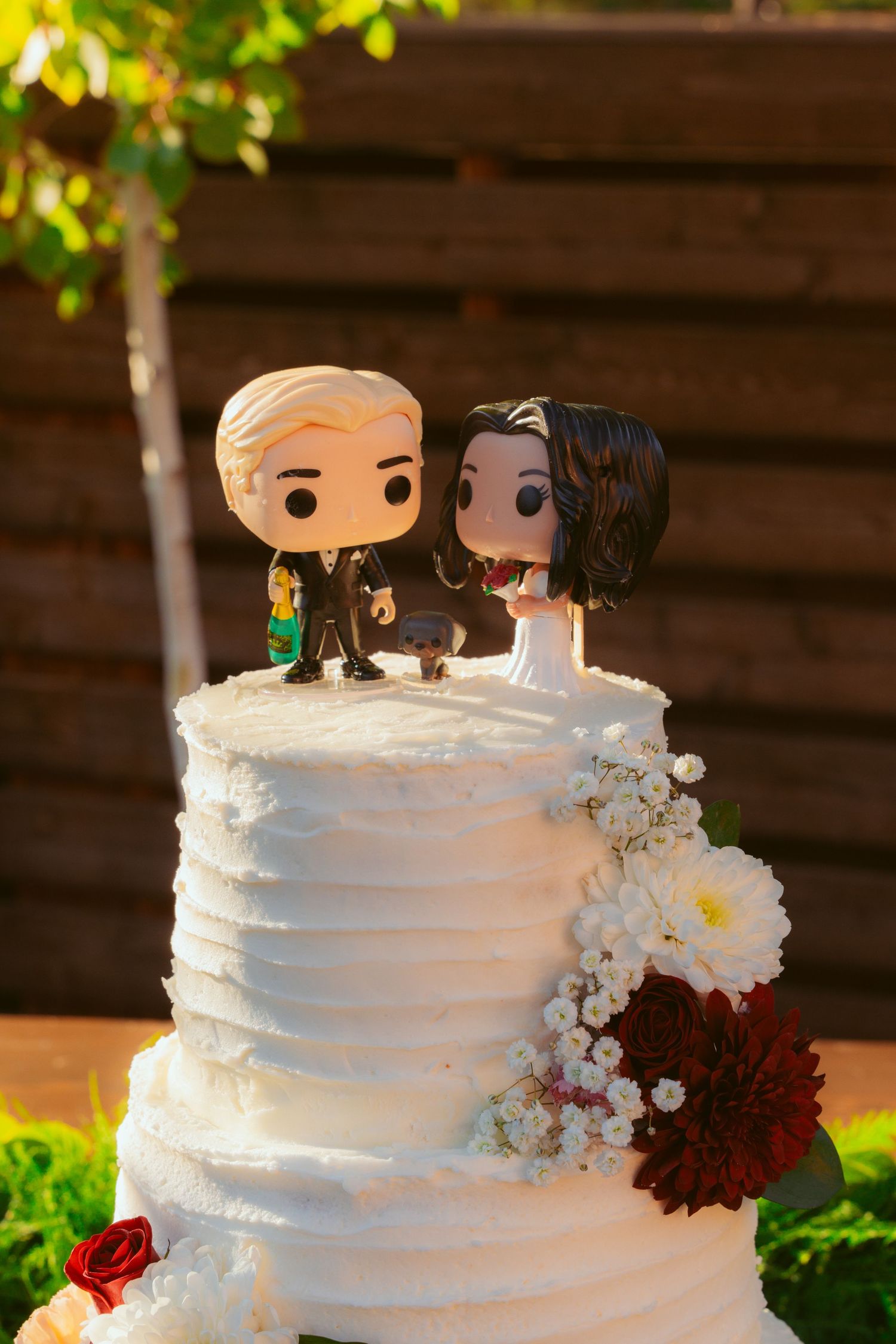 White wedding cake topped with decorative figurines and burgundy flowers on a rustic wooden display.