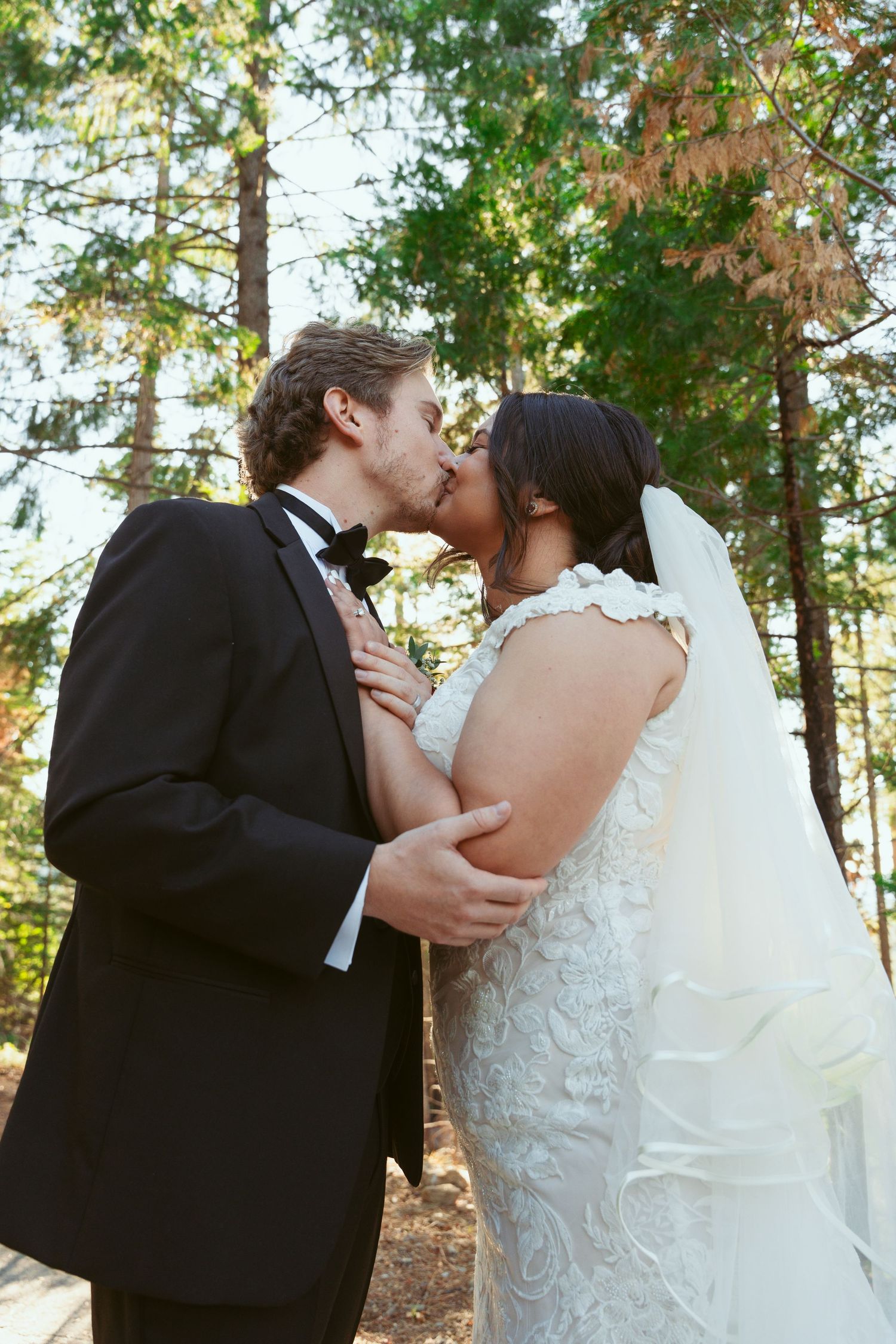 A romantic wedding kiss between newlyweds in a sunlit forest setting.